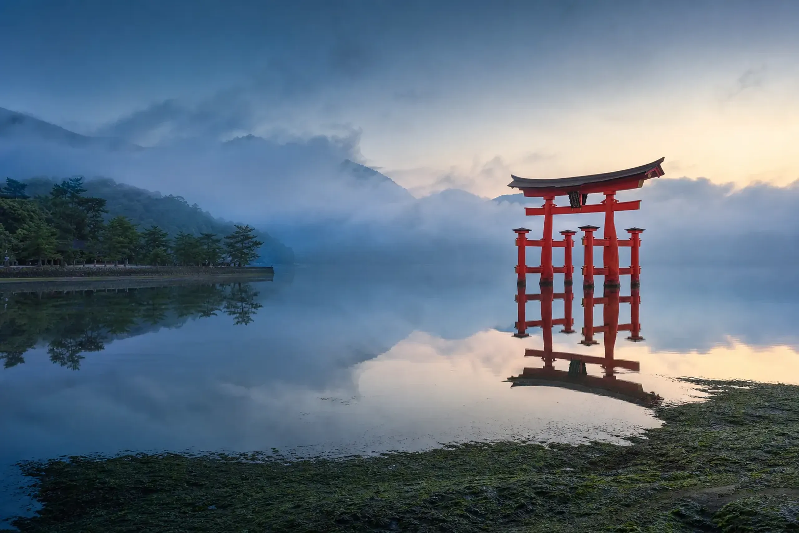 The crimson floating torii gate of Itsukushima Shrine at dawn on Miyajima island, standing in the still shallow waters of the Seto Inland Sea, mist over the surrounding forested hills