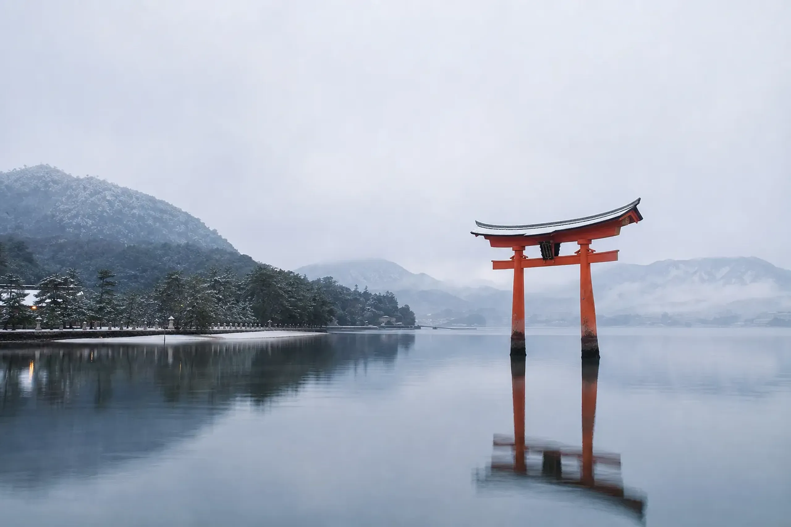 The crimson floating torii gate of Itsukushima Shrine at dawn on Miyajima island, standing in the still shallow waters of the Seto Inland Sea, mist over the surrounding forested hills