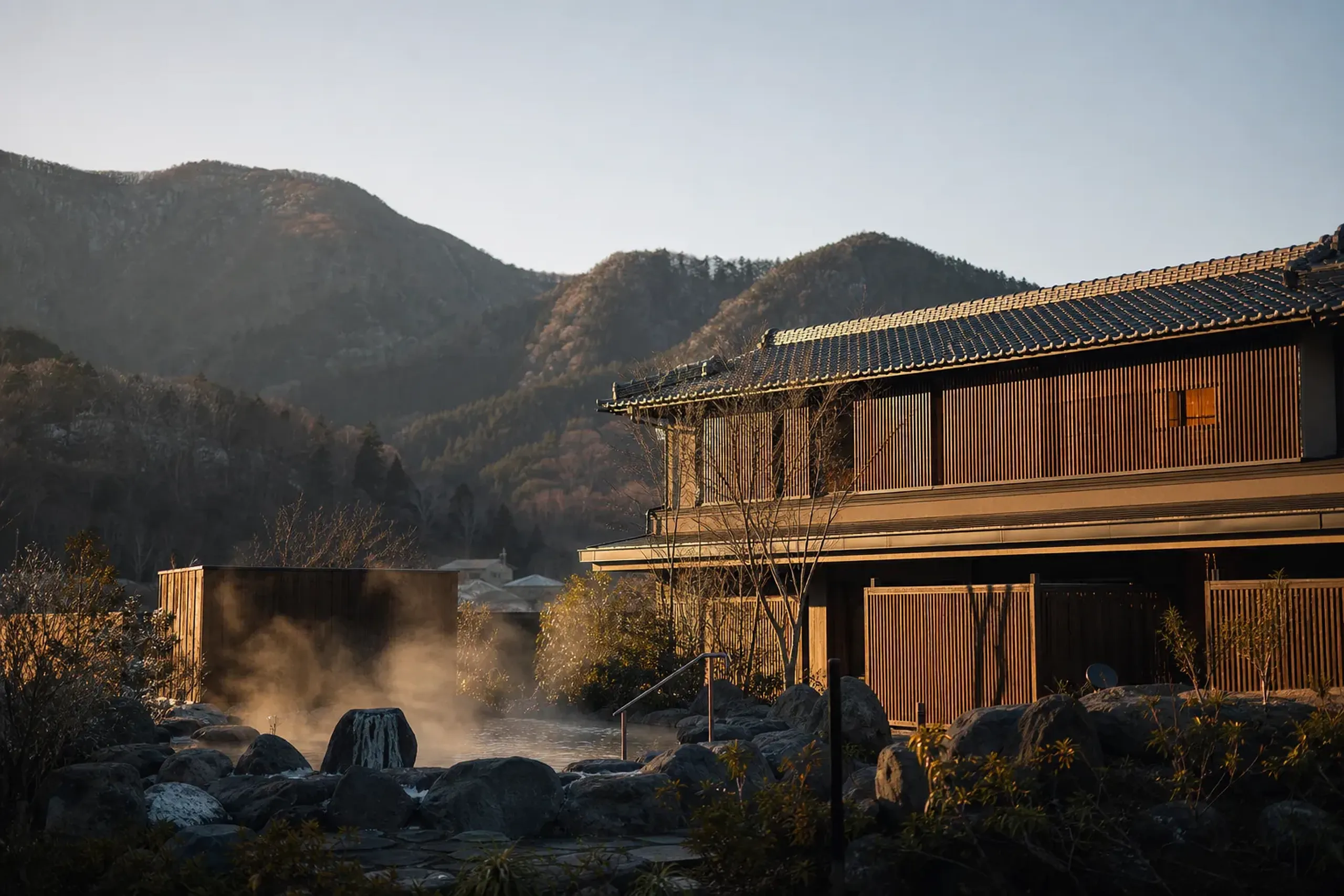 The design-forward exterior of the newly opened KAI Kusatsu onsen ryokan by Hoshino Resorts, with steam rising from a hot-spring bath against the mountain backdrop of Kusatsu