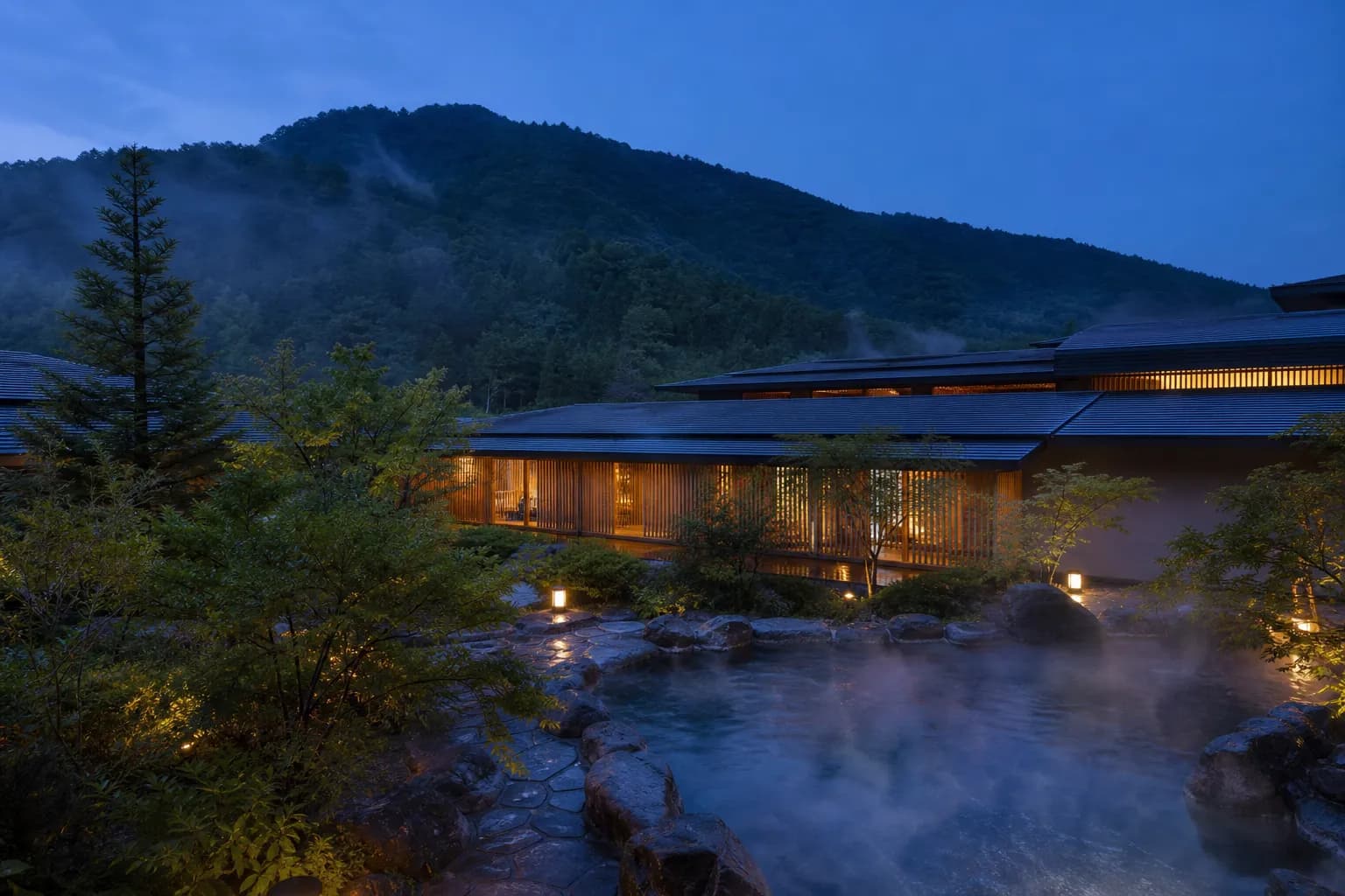 The design-forward exterior of the newly opened KAI Kusatsu onsen ryokan by Hoshino Resorts, with steam rising from a hot-spring bath against the mountain backdrop of Kusatsu