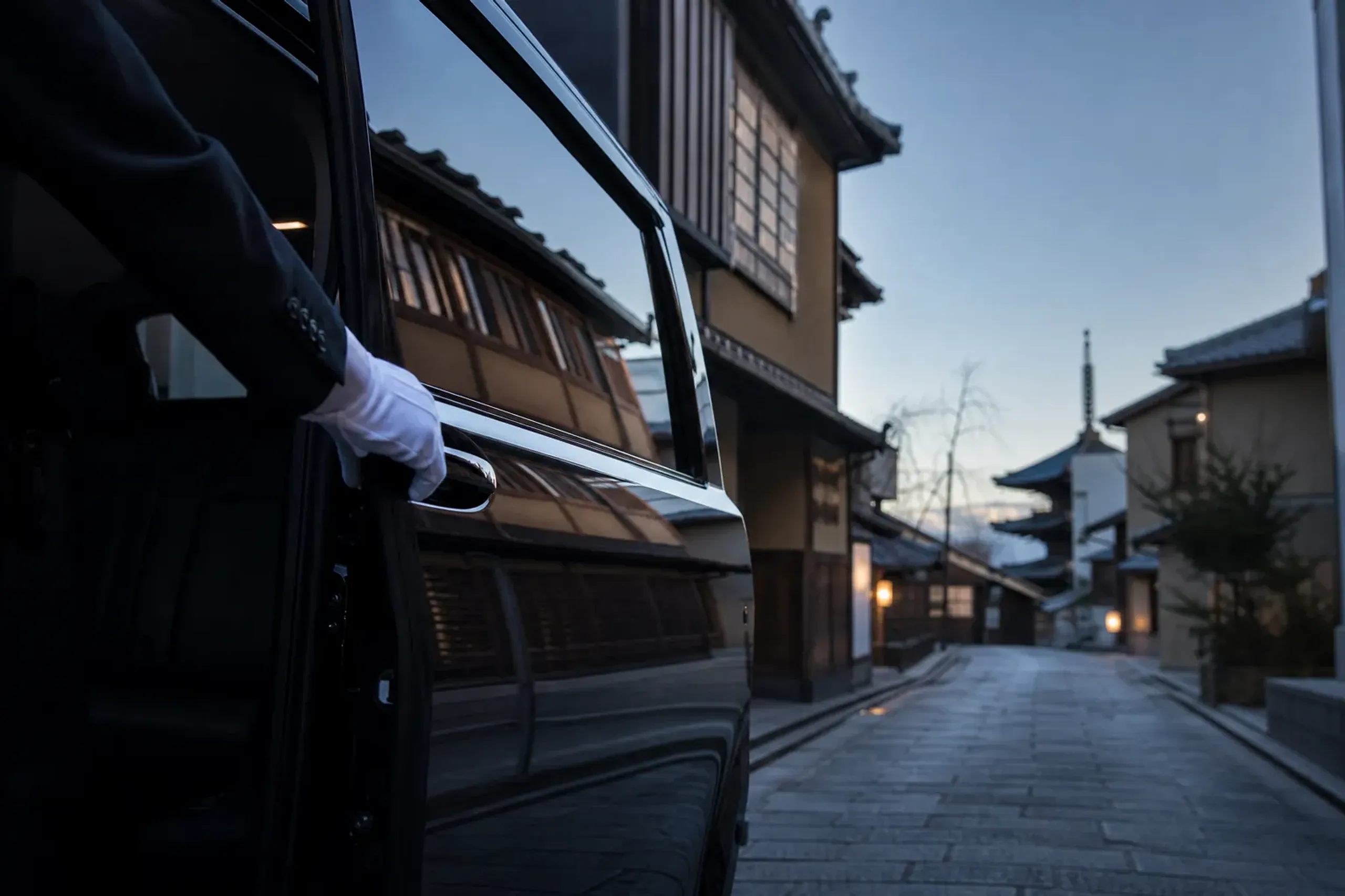 A chauffeur's white-gloved hand opening the rear door of a Toyota Alphard at a Kyoto machiya street corner at dawn