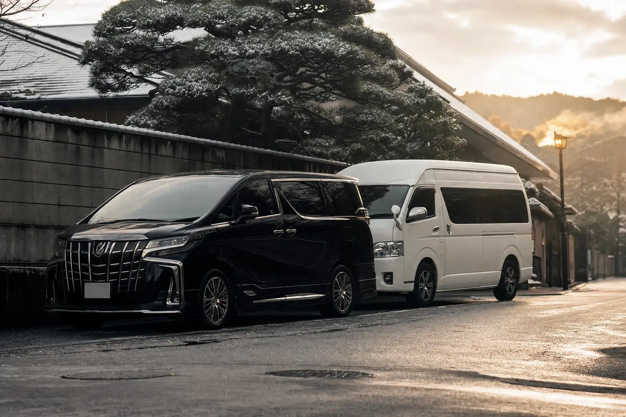 A Toyota Alphard and a larger Toyota HiAce parked side by side on a quiet Japanese street, showcasing their contrasting sizes