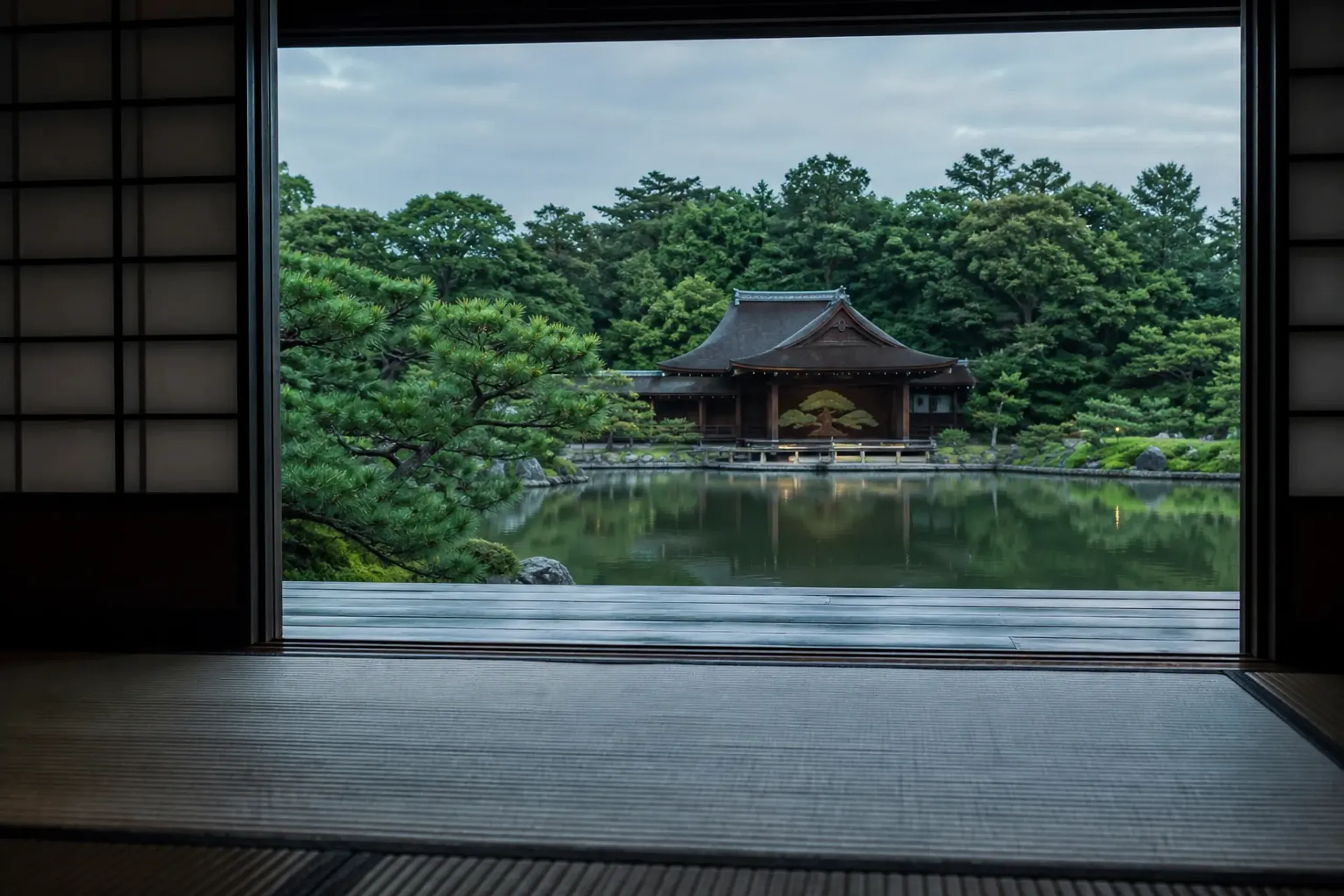 A tatami-floored guest room at Asaba Ryokan with shoji screens open onto an engawa veranda, framing a view across the pond to the Gekkeiden Noh stage