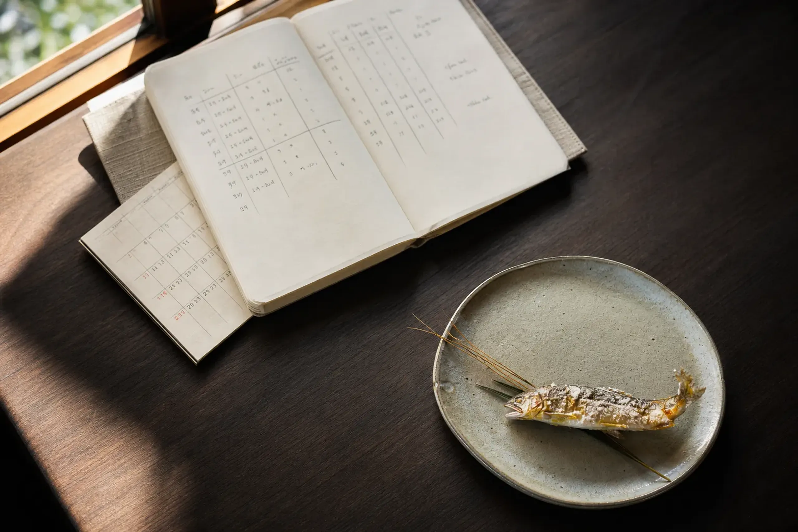 Notebook and calendar used to plan ukai season dates and dinner timing, paired with a small seasonal dish