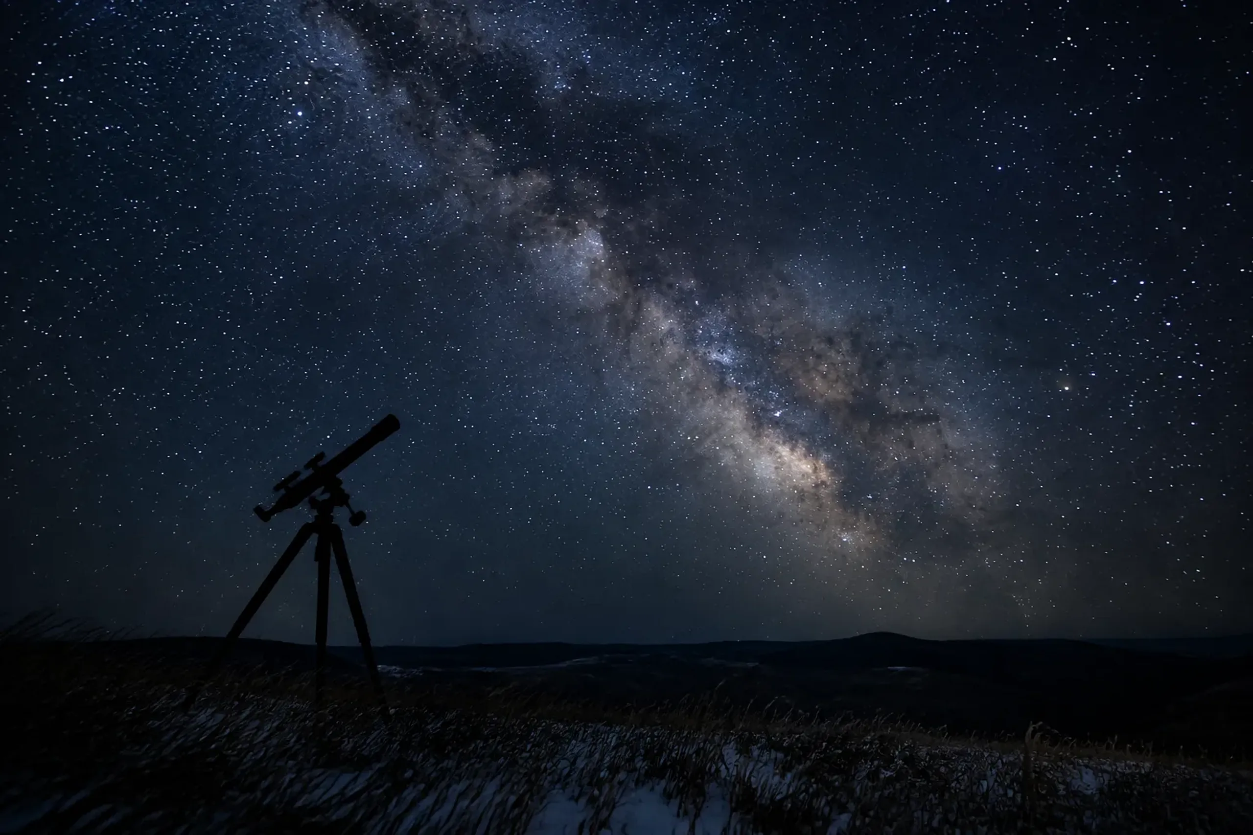 A telescope mounted on a tripod pointed toward the Milky Way above the wind-swept dark hills surrounding Shosanbetsu Astronomical Observatory in Hokkaido