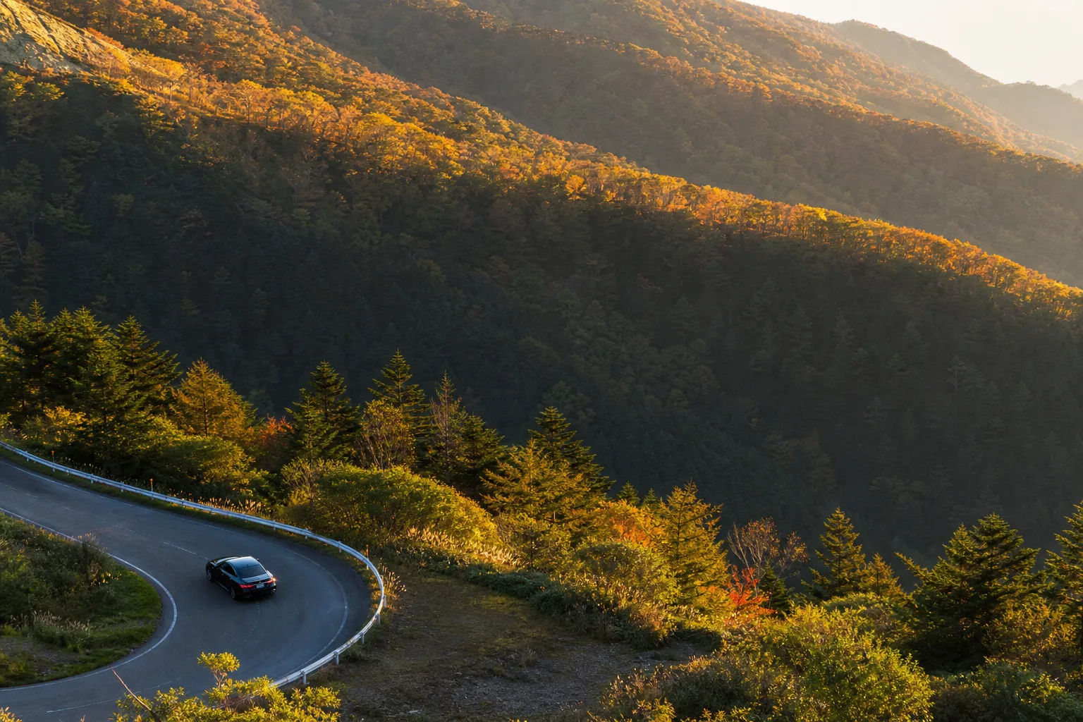 A luxury vehicle driving on a winding mountain pass in northern Japan, where high-altitude autumn golds contrast sharply with the deep green forests of the lower valleys.