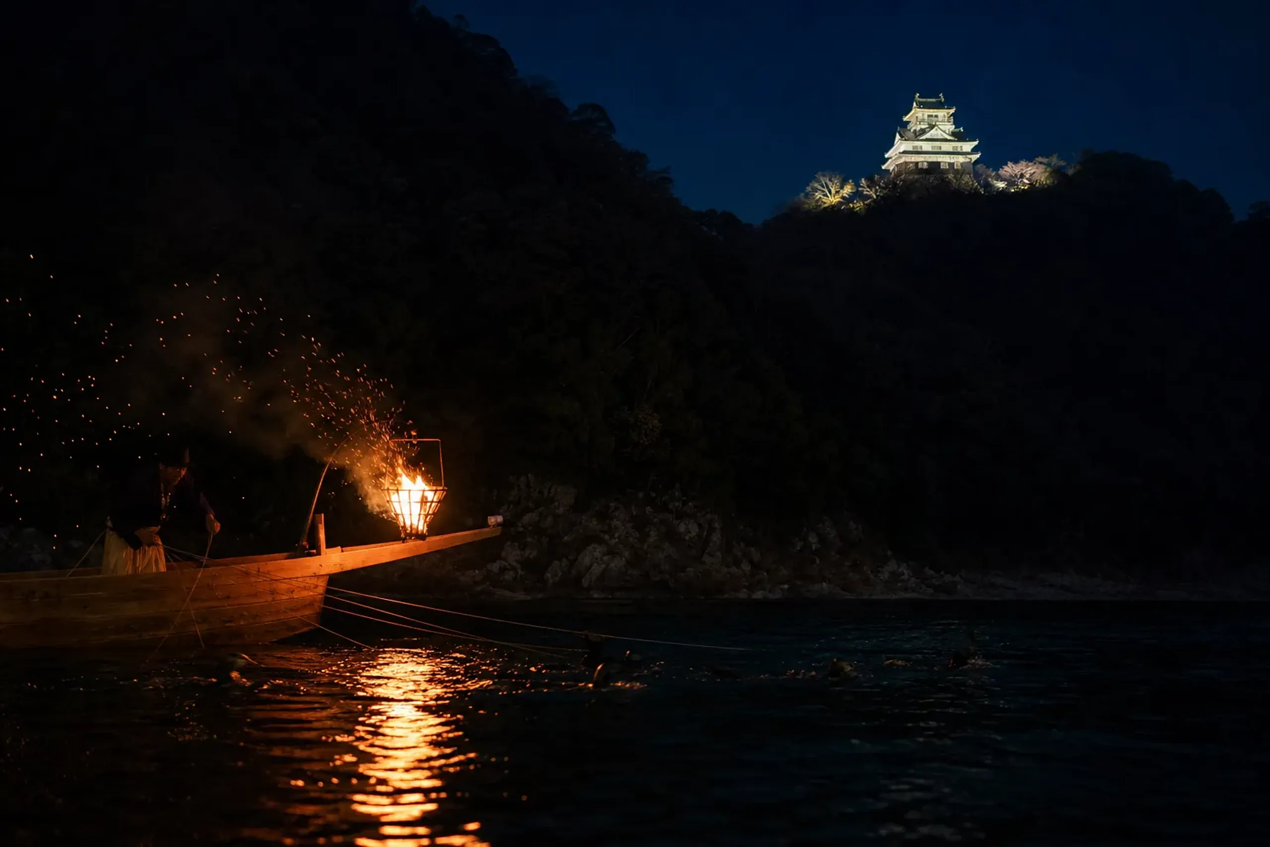 A cormorant fishing boat on Gifu's Nagara River at night with iron fire baskets blazing over the bow, Gifu Castle illuminated on the mountain above the dark water