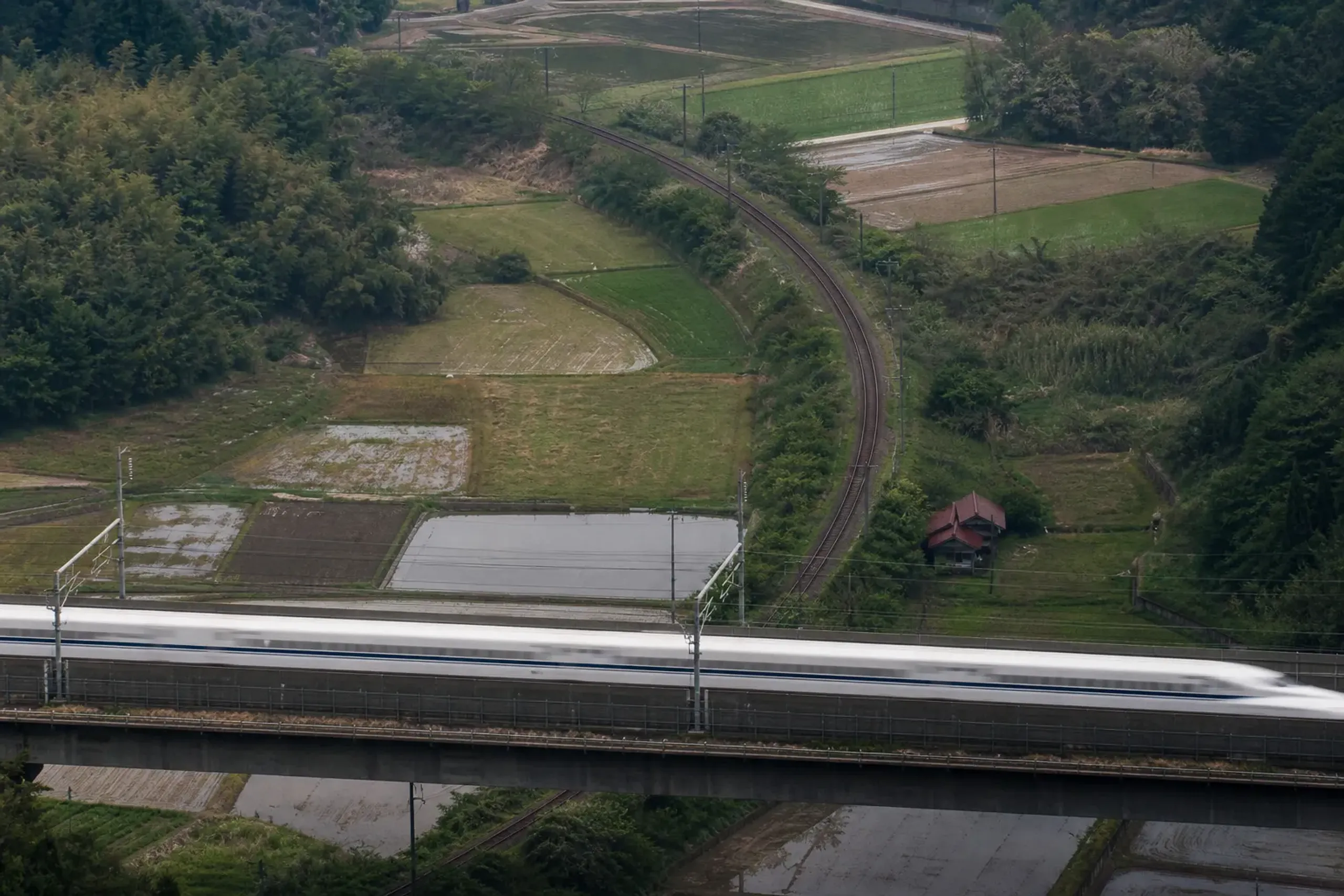a shinkansen bullet train streaking past on the Tokyo-Kyoto-Osaka spine while, in the distance, a single-track rural rail line winds through empty countryside, illustrating how rail thins beyond the G
