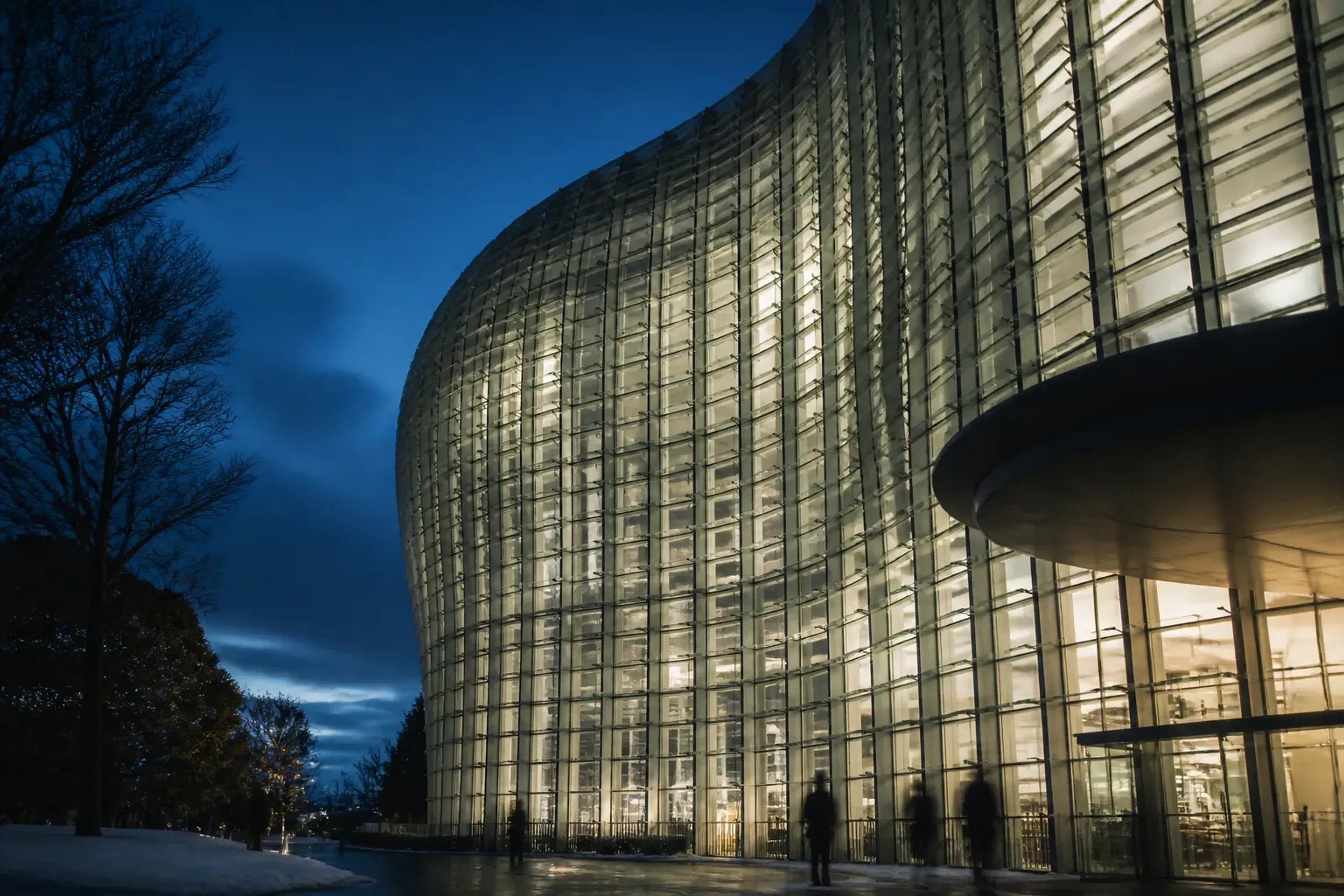 Exterior architectural shot of The National Art Center, Tokyo (Kurokawa’s curved glass facade) photographed at dusk to anchor the 2026 viewing route context, with no readable signage and no identifiab