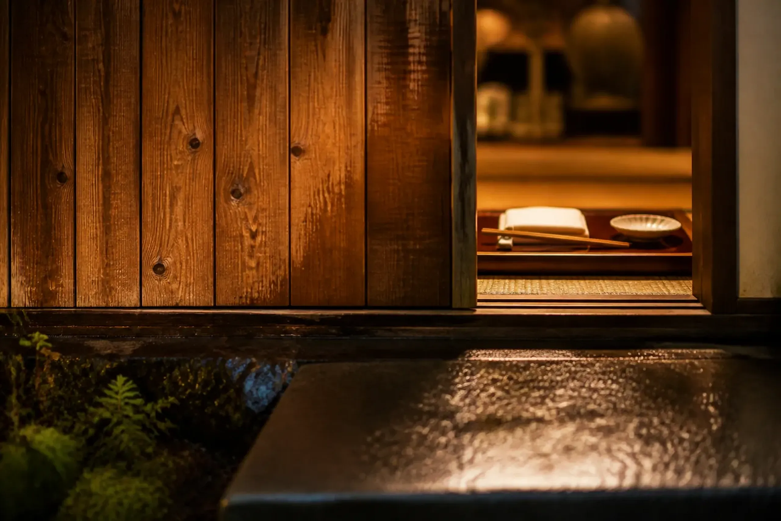 A close-up, dusk-lit view of a cedar-wood entryway at a private Japanese house with warm lantern light raking across the cedar grain and an untouched, fully set dining place setting visible just insid