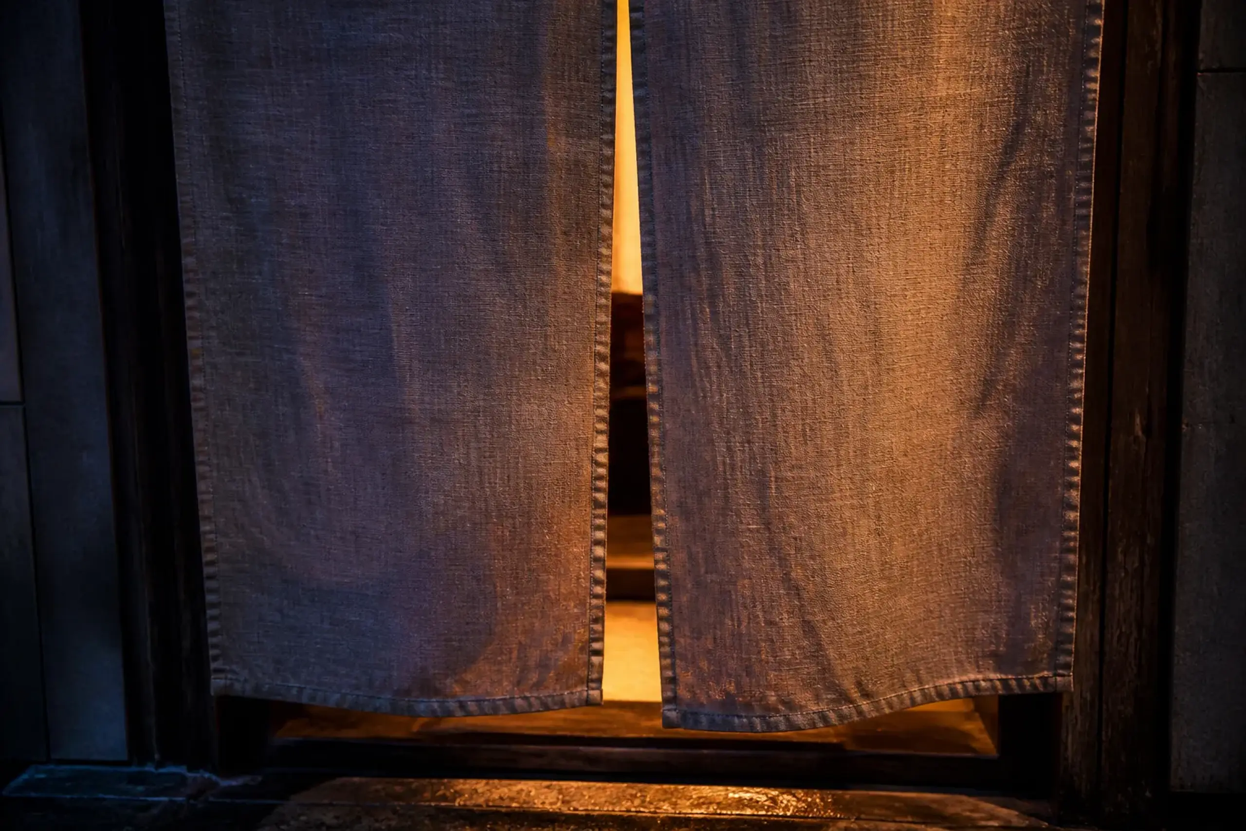 A tight composition of a plain noren (unmarked fabric doorway curtain) at the entrance to a small street-side establishment, photographed at twilight with soft spill light beyond the curtain—emphasizi
