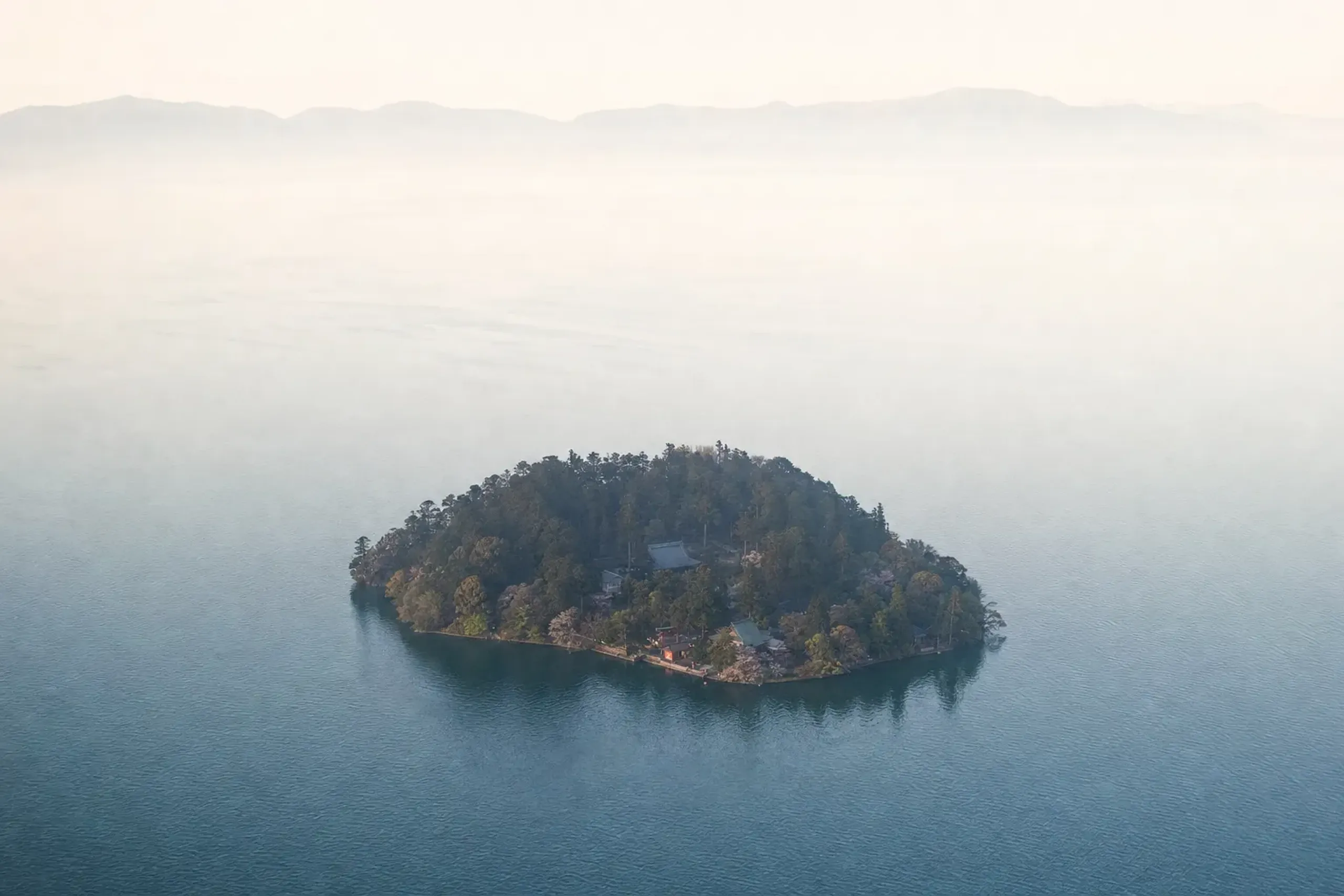 Aerial view of the small sacred island of Chikubushima rising densely wooded from Lake Biwa, its shrine rooftops nestled among the trees
