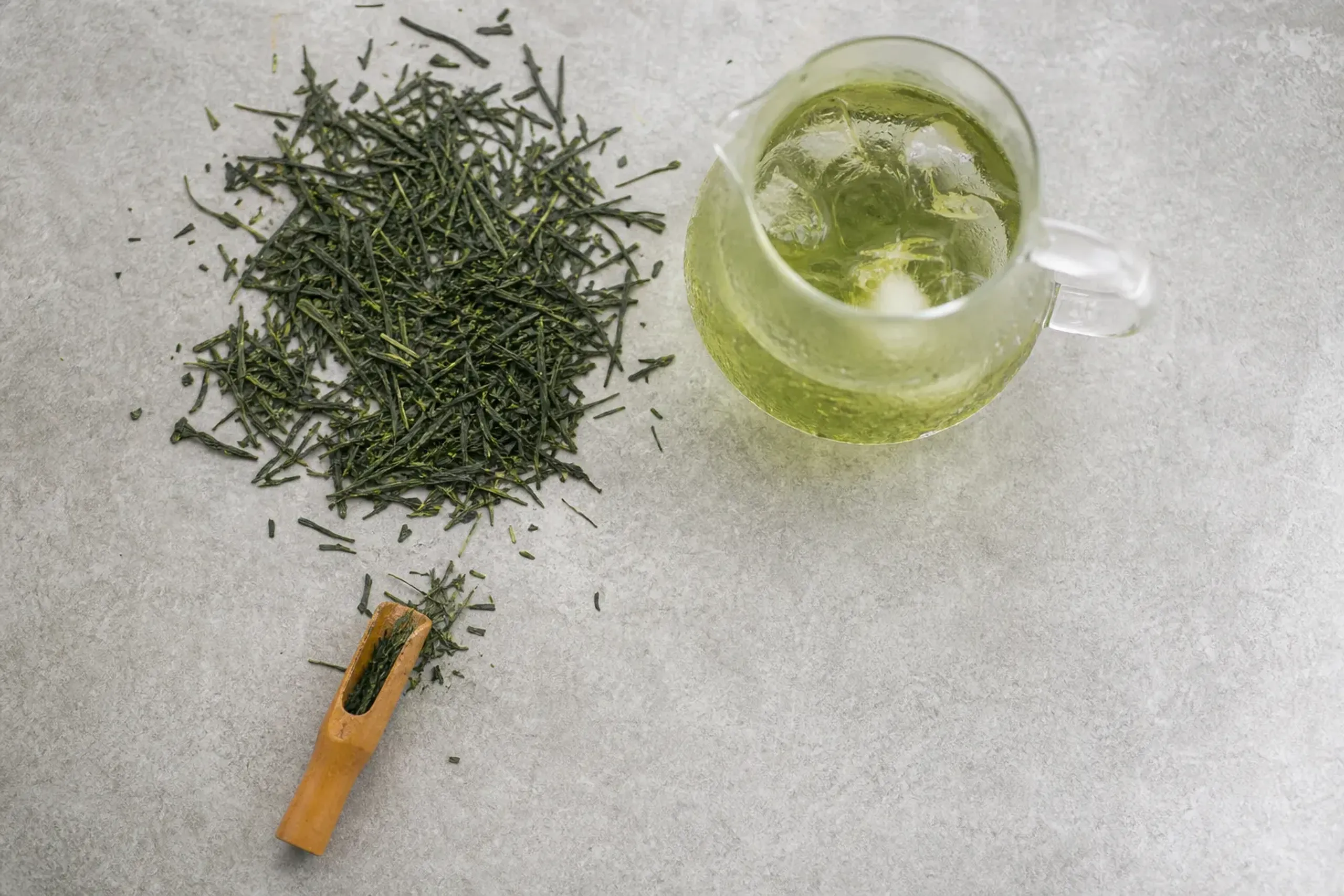 Loose sencha leaves, a glass pitcher of cold brew, and ice cubes arranged for home brewing