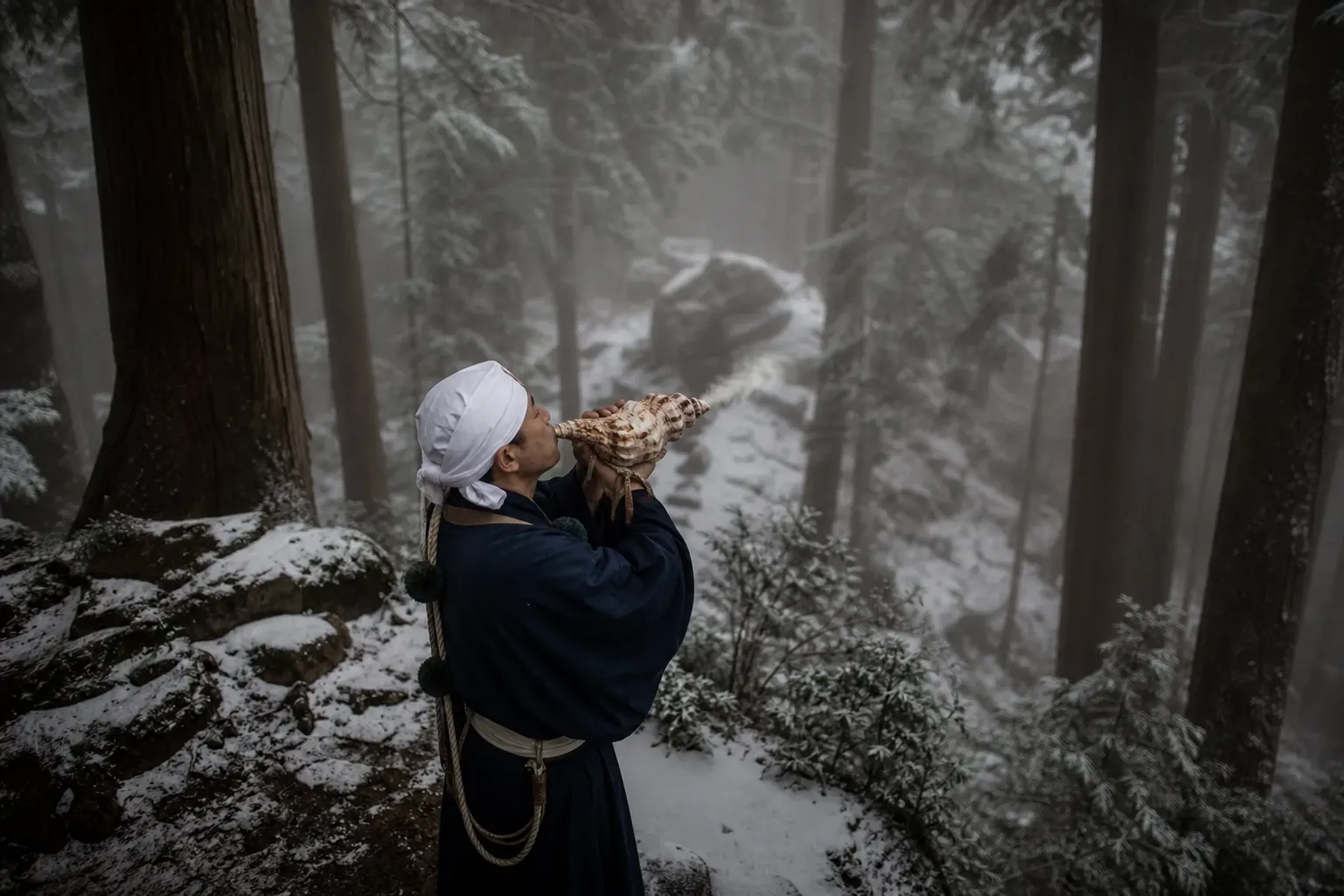 Man in traditional blue robes and a headwrap holds and kisses a large conch shell in a forest.