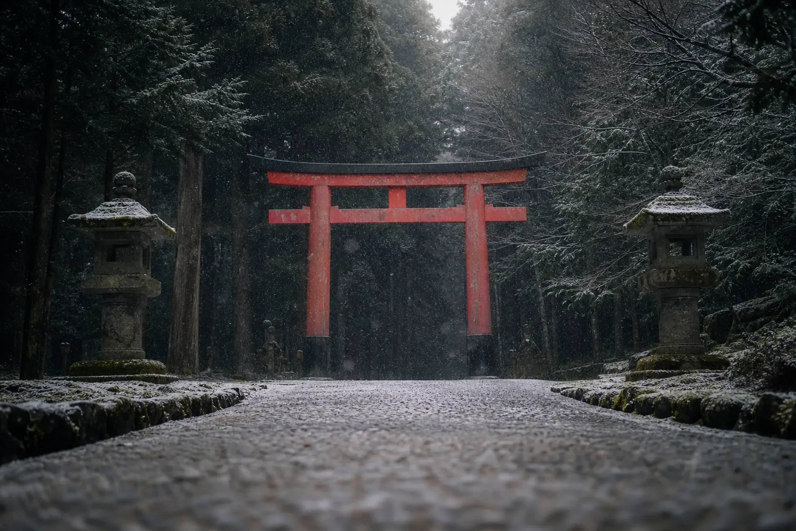Red torii gate at shrine entrance along a gravel path, with stone lanterns and lush green trees surrounding it.