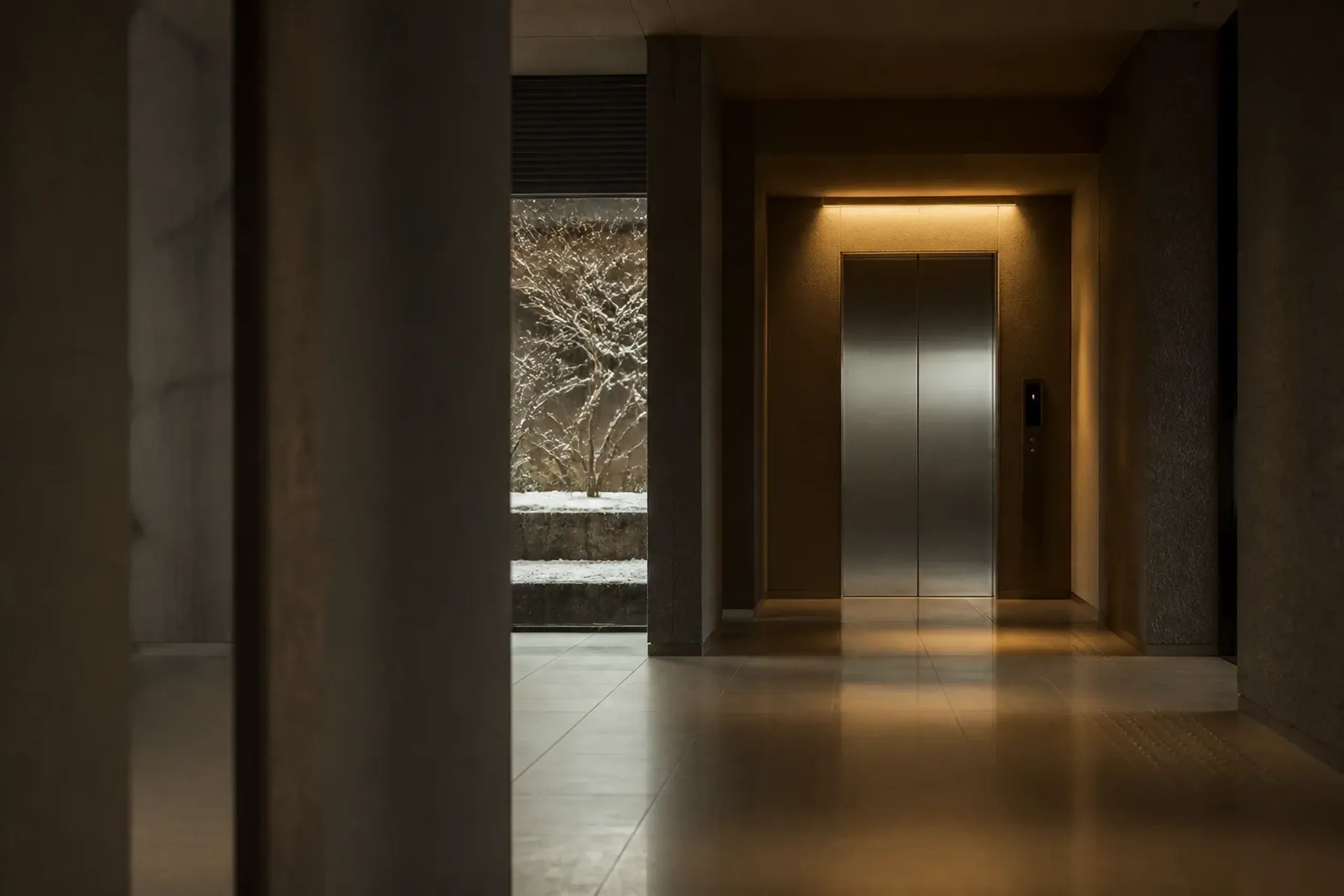 An empty hotel lift bank with brushed-steel doors and a softly lit threshold, viewed from an adjacent quiet corridor at an off-peak hour