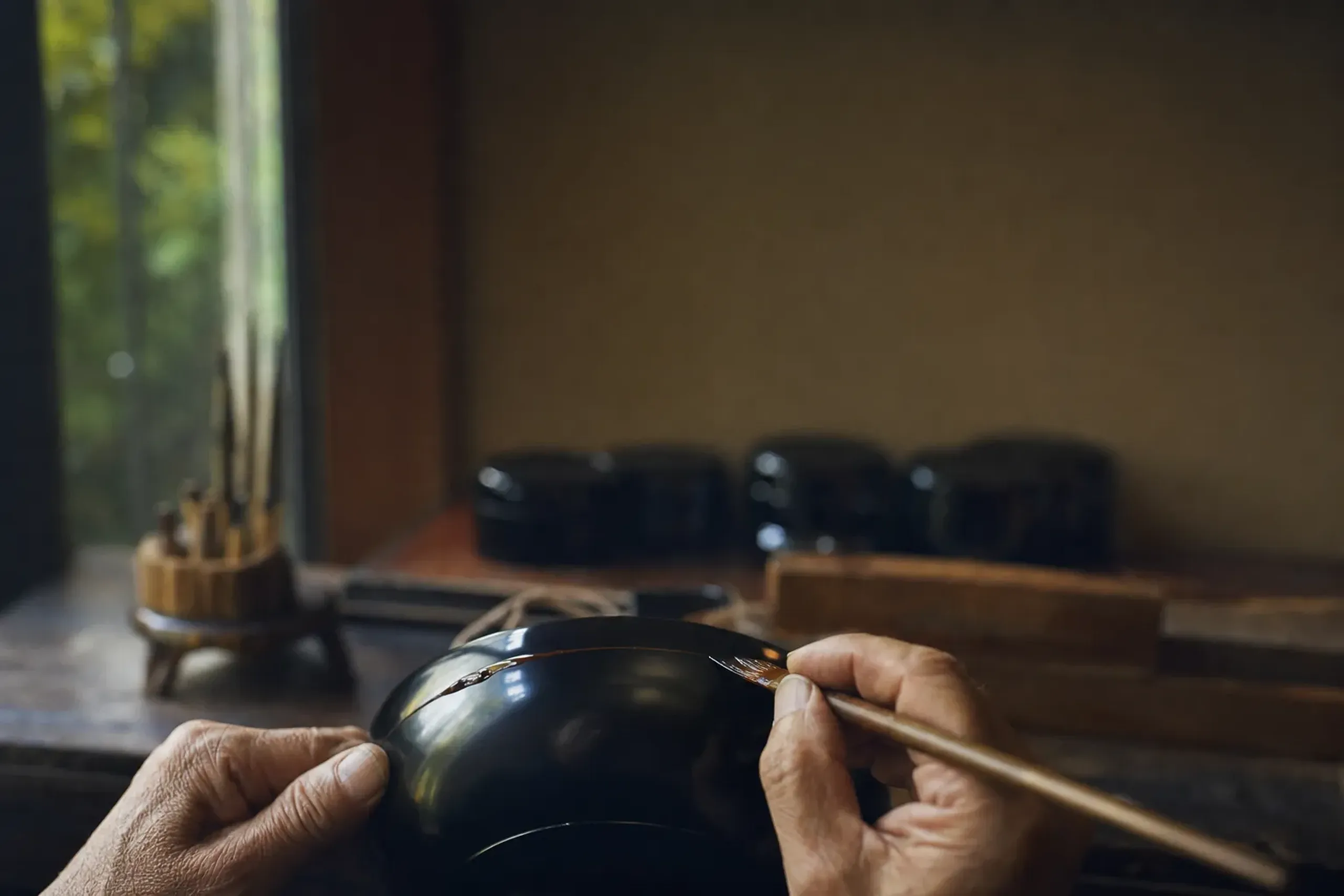 the hands of a master lacquerware shokunin applying urushi lacquer with a fine brush to a glossy black bowl in a Kyoto artisan workshop, tools and finished pieces arranged on the bench