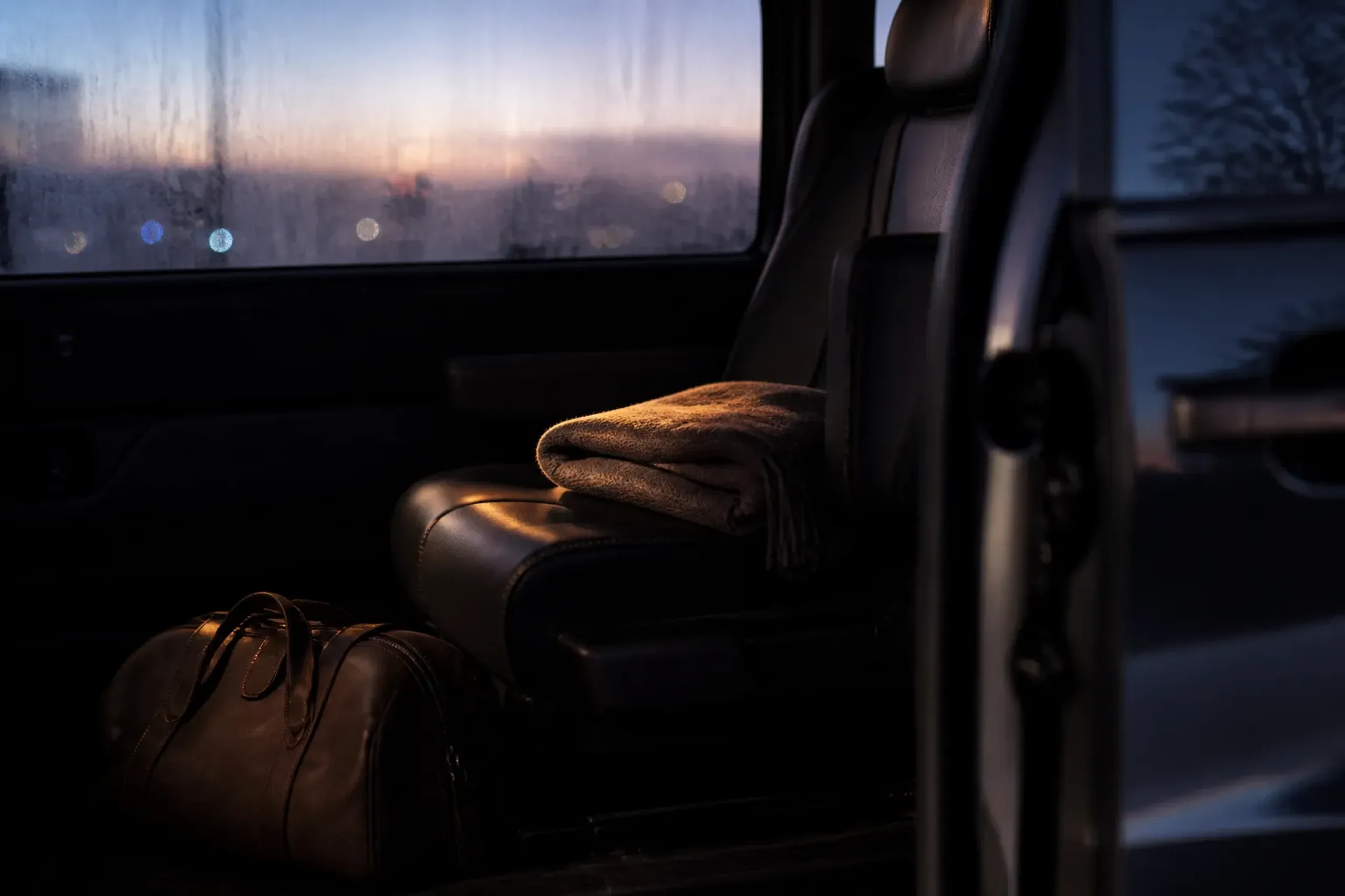 Interior of a chauffeured luxury minivan at dawn with a small travel bag and city lights outside