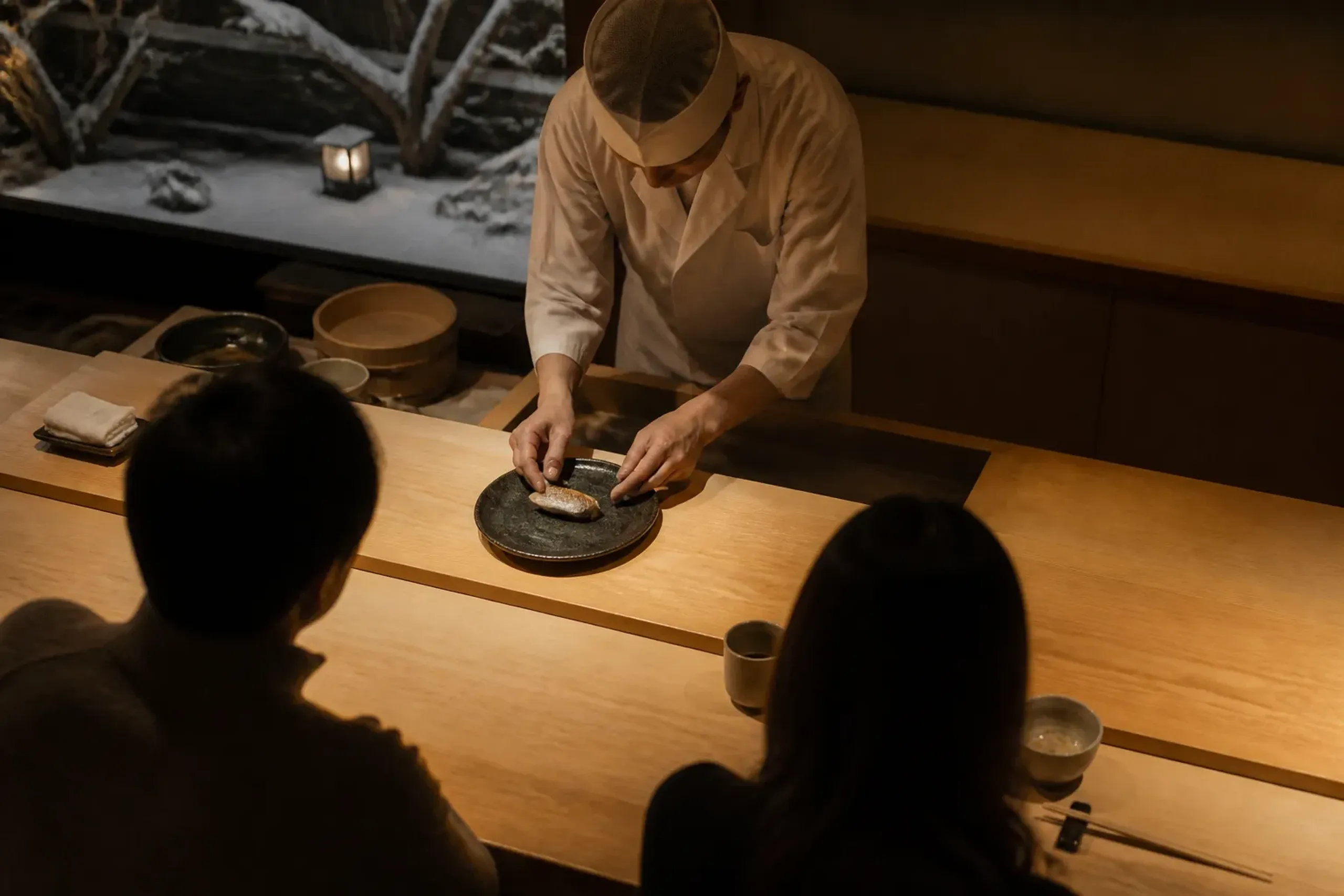 Warmly lit intimate Tokyo restaurant counter at night with a chef plating food
