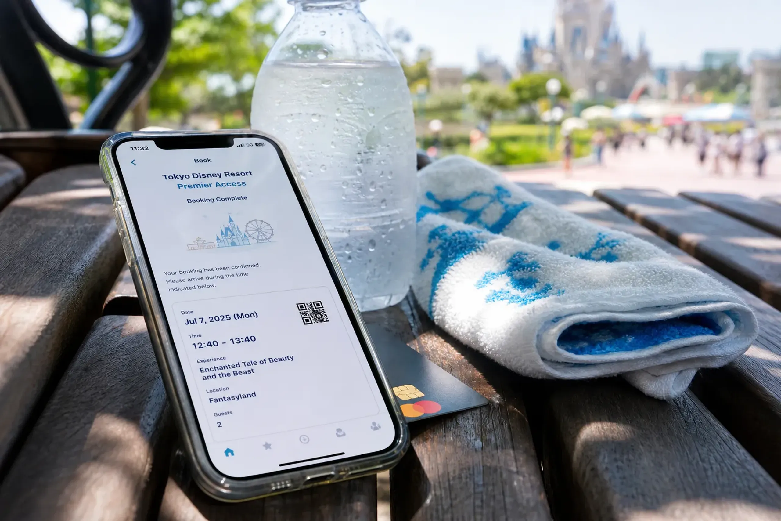 A single smartphone with a payment card resting on a shaded park bench beside a cooling towel and water bottle at Tokyo Disney Resort