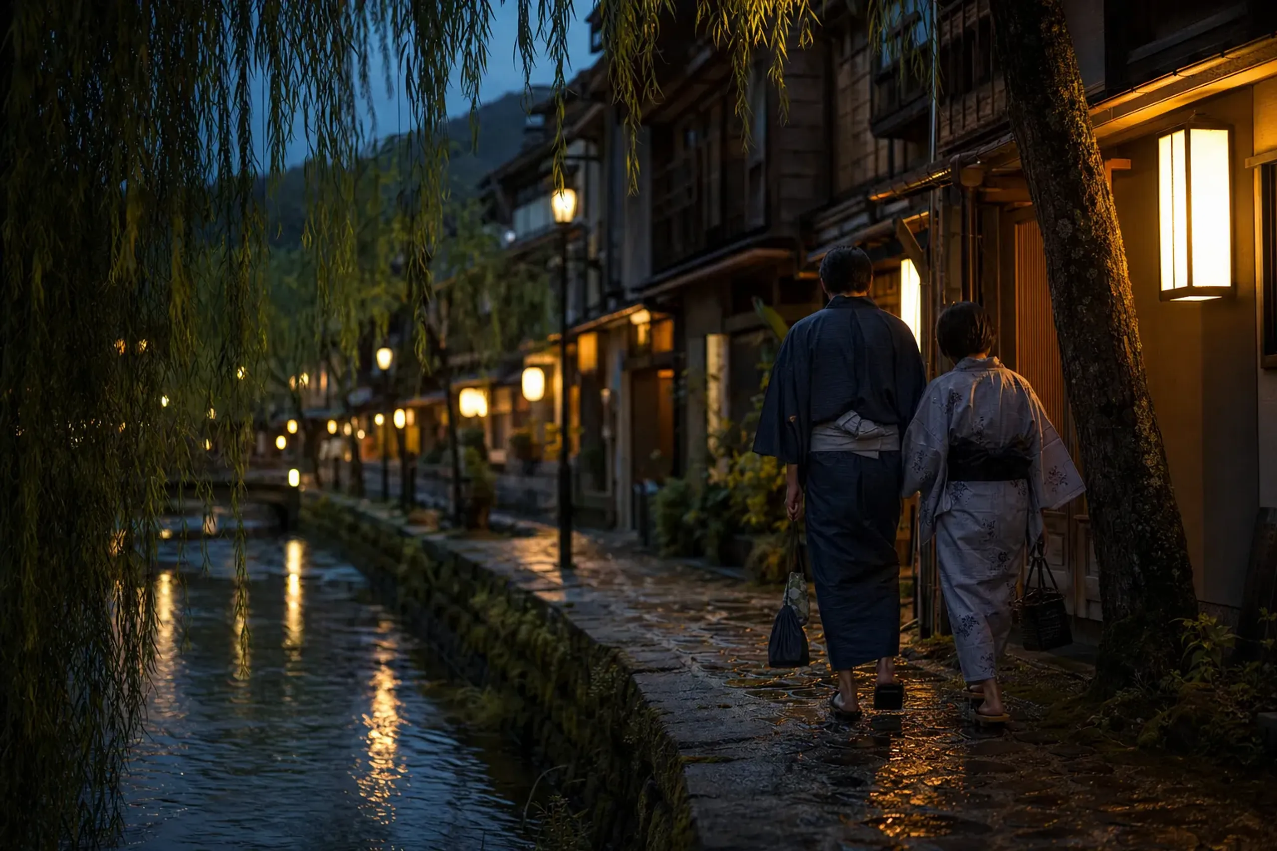 Guests in yukata and geta walking a willow-lined lane between bathhouses in Kinosaki Onsen at dusk