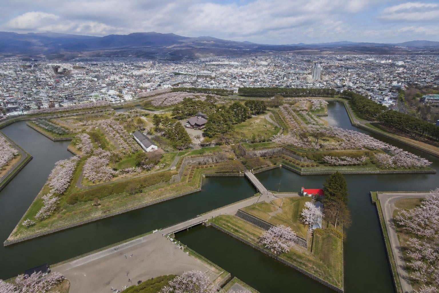Aerial view of a historic Japanese castle garden with cherry blossoms, moats, and stone walls overlooking a city skyline and distant mountains.