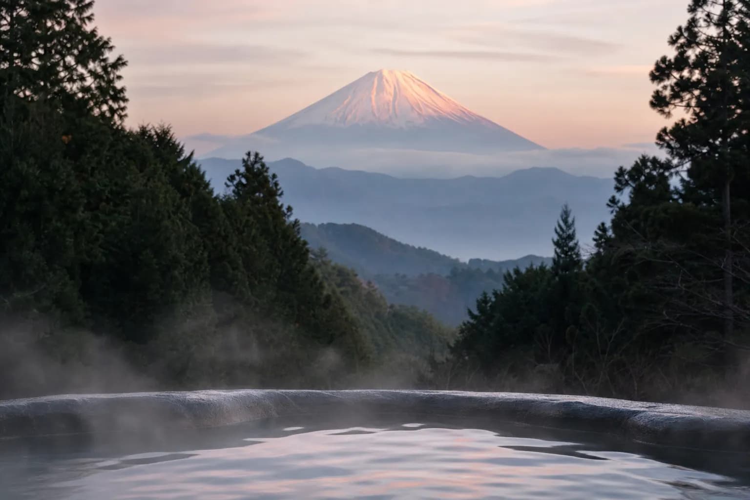 A steaming open-air onsen among misty forested hills with Mount Fuji rising behind, in Hakone