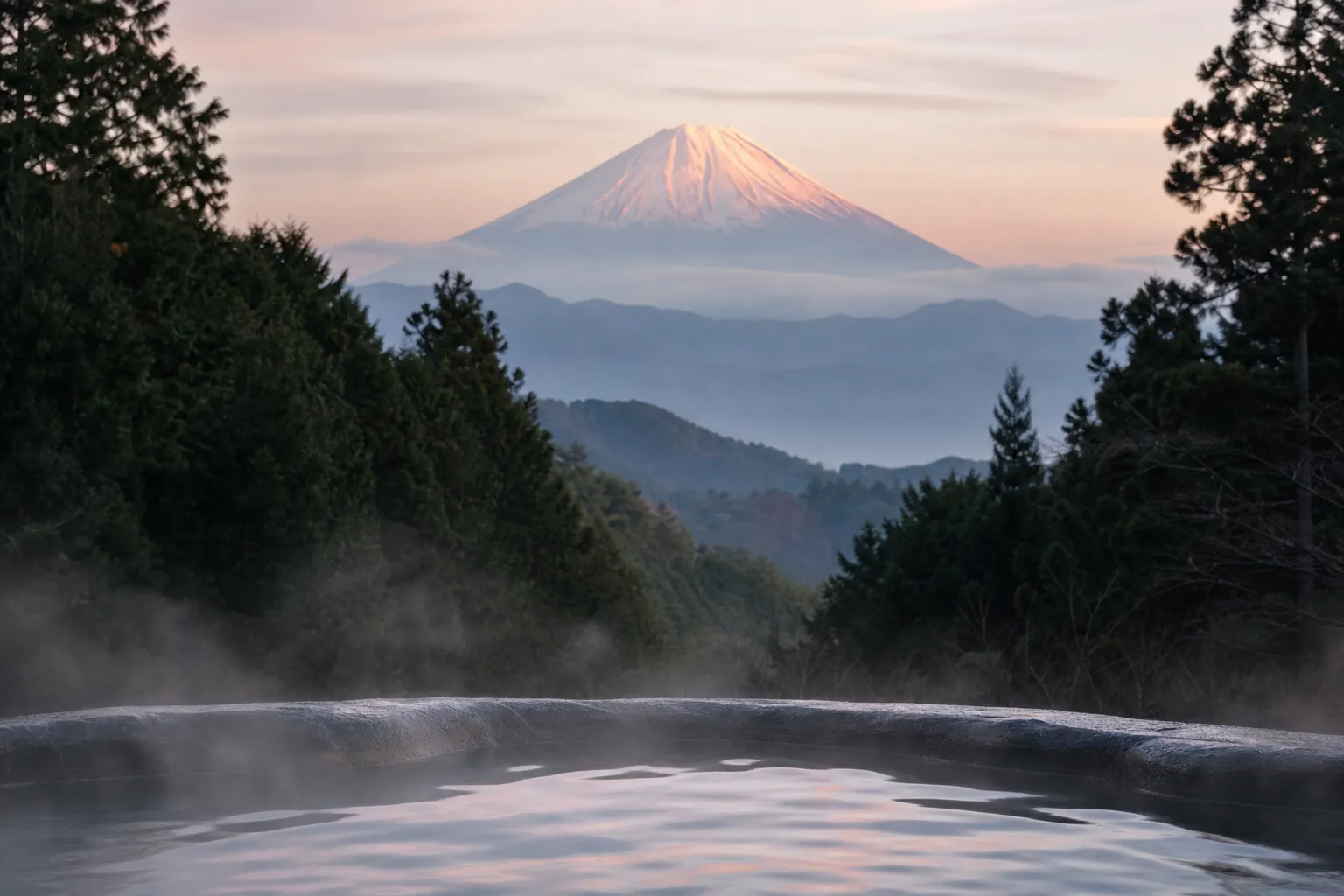 A steaming open-air onsen among misty forested hills with Mount Fuji rising behind, in Hakone