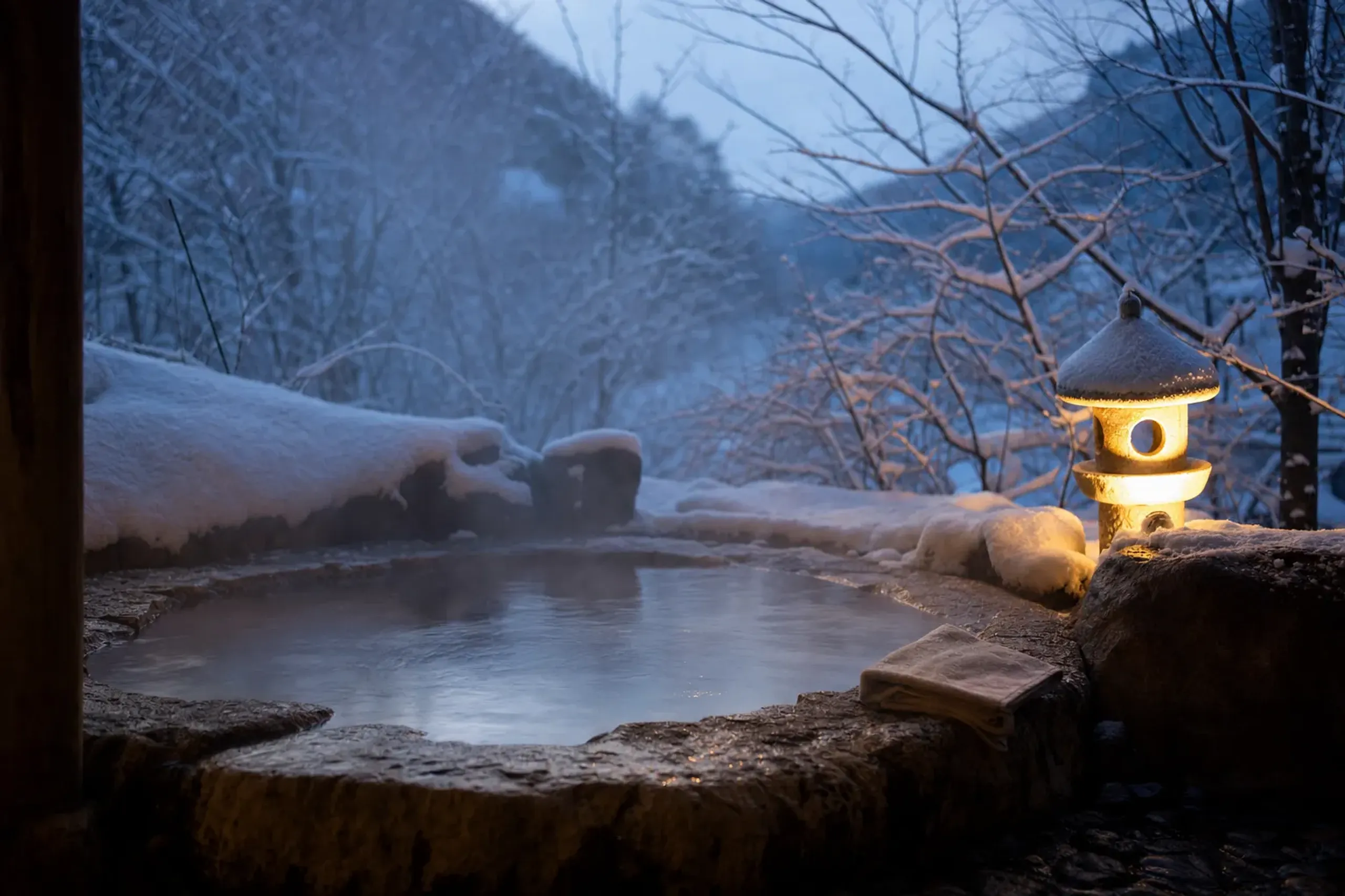 Open-air onsen bath surrounded by snow at dusk with steam rising