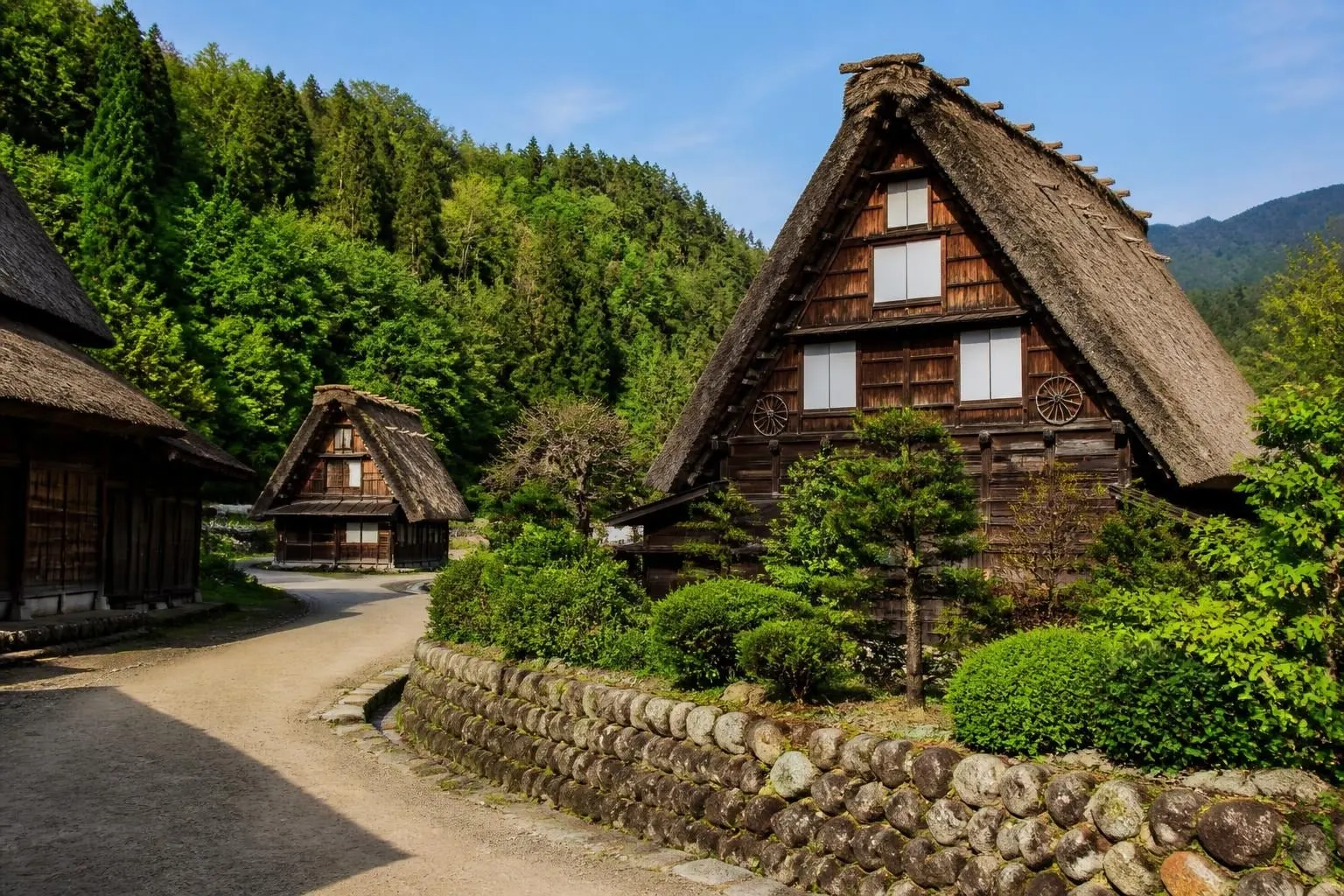 Traditional thatched-roof houses along a gravel path in a green, forested village setting with a stone retaining wall.|Informative scene in rural Japan showing gassho-style houses and lush greenery