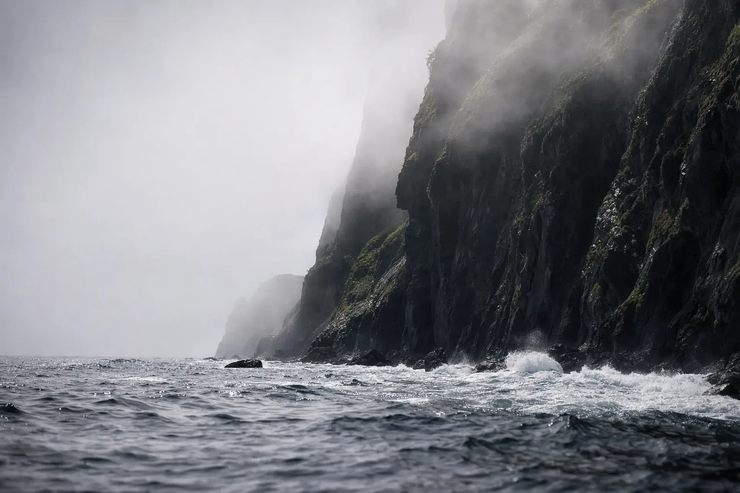 The wild, fog-shrouded cliffs of the Shiretoko Peninsula jutting into the rough, gray waters of the Sea of Okhotsk under shifting summer weather