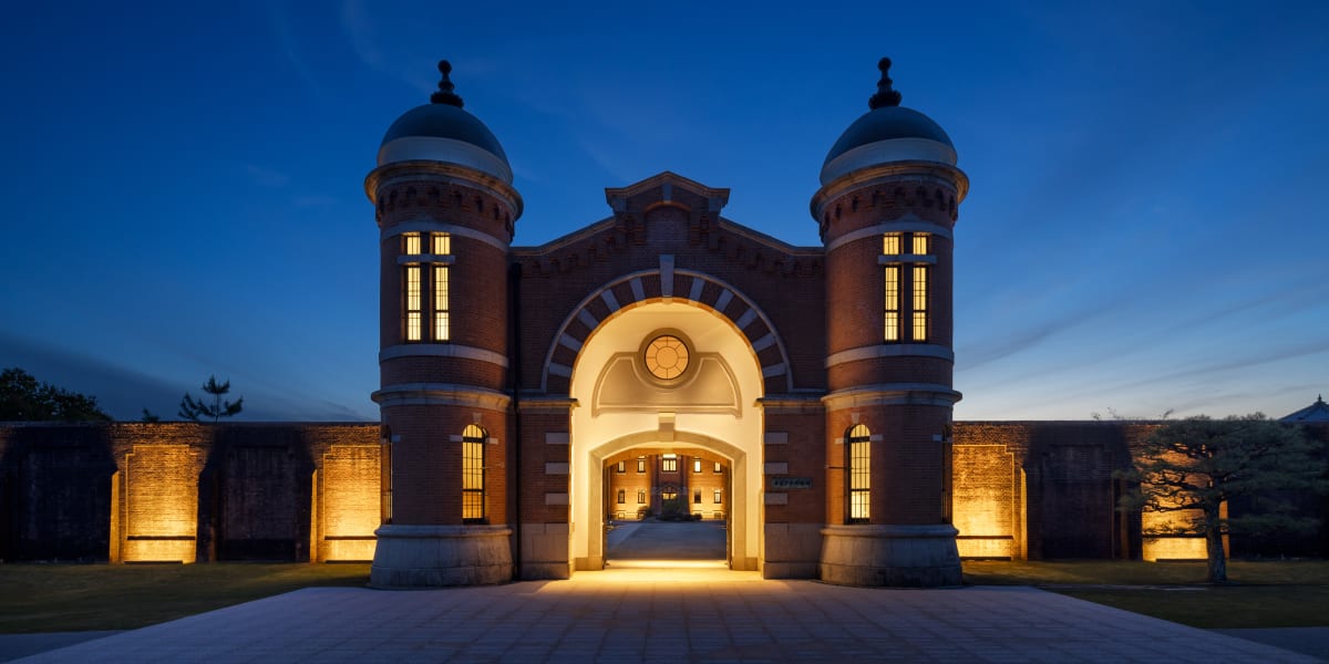 A symmetrical, Meiji-era red brick gatehouse at twilight, featuring two grand cylindrical towers with domes flanking a large, brightly illuminated central archway. Warm architectural uplighting highlights the brick texture and surrounding walls against a deep blue evening sky.