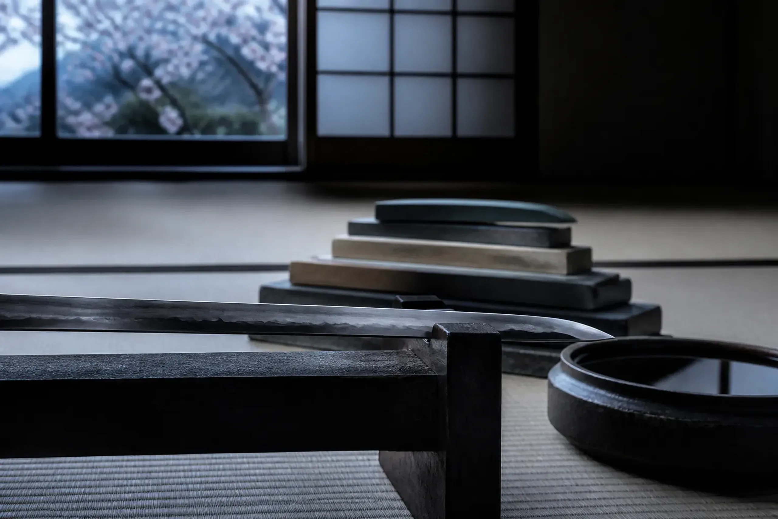A togishi's polishing studio interior in total silence, an antique blade resting on a wooden work stand beside stacked whetstones, with no forge or fire present