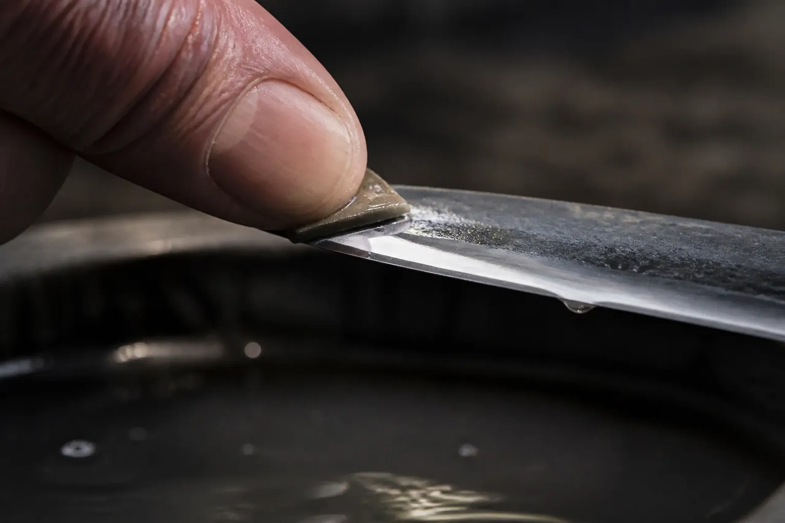Paper-thin uchigumori hazuya and jizuya stone fragments affixed with lacquer to a polisher's thumb, rubbing across a wet blade section above a water basin