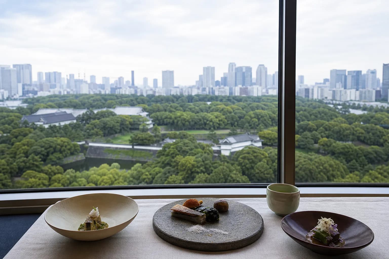 Table set with three plated dishes and a cup of tea in a-windowed restaurant overlooking a green park and city skyline far in the distance, daytime view