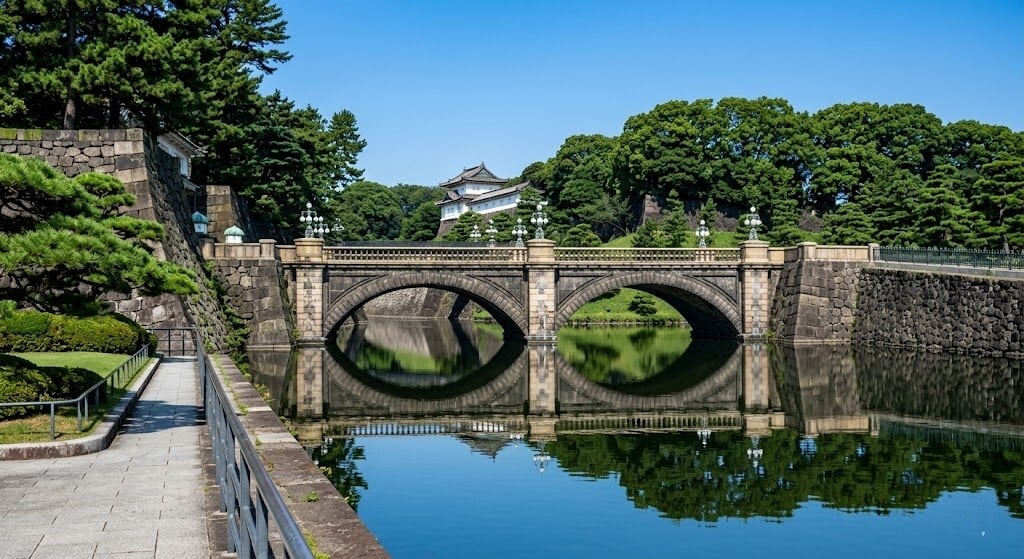 Stone arch bridge spanning a calm moat, with reflected arches and surrounding stone walls and trees.