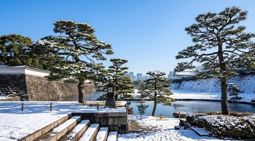 Snow-covered Japanese castle grounds with stone walls, pine trees, and a tranquil moat under a blue winter sky.