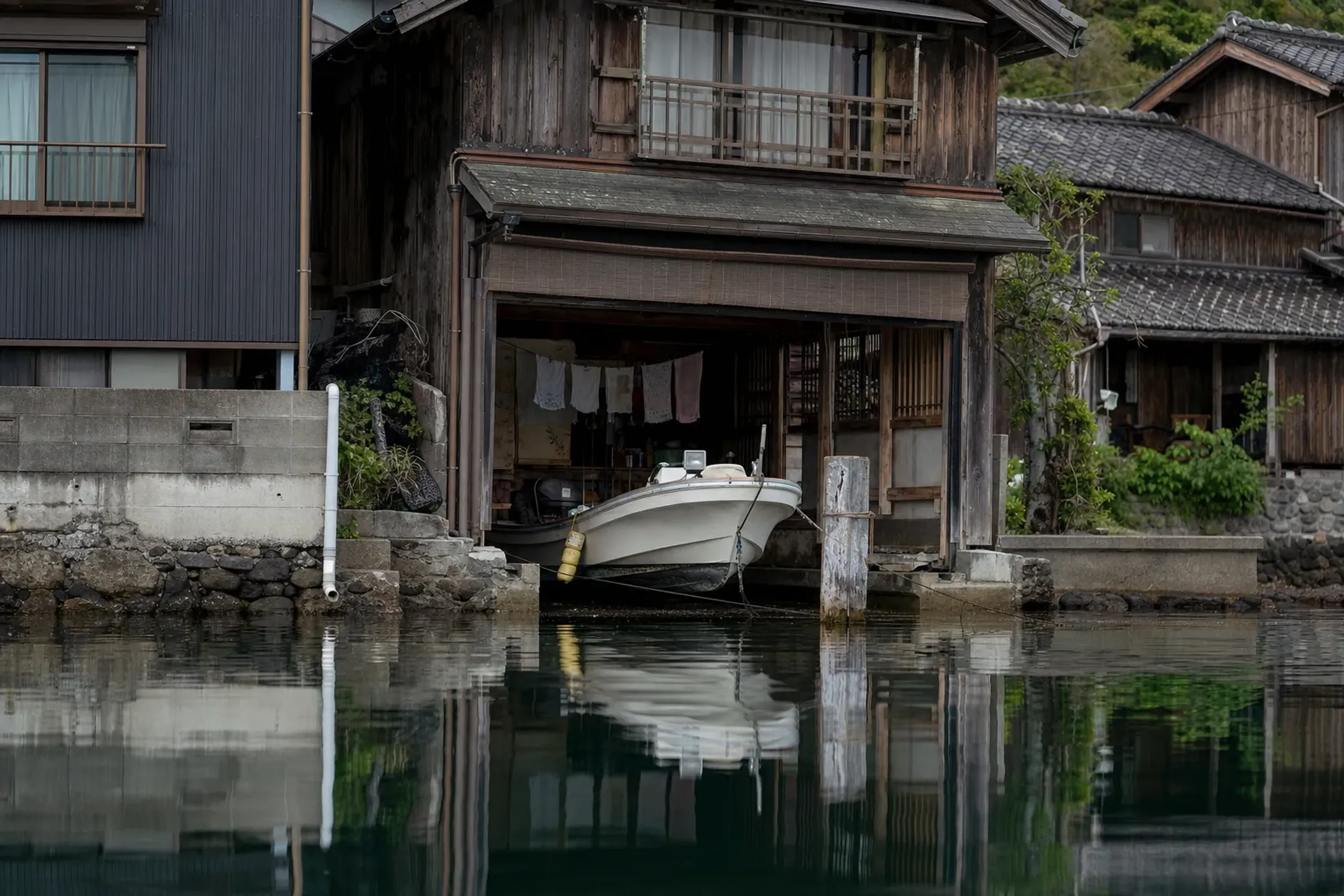 A modern fibreglass work boat moored in the water in front of an Ine funaya, with laundry hanging in the open ground-floor bay