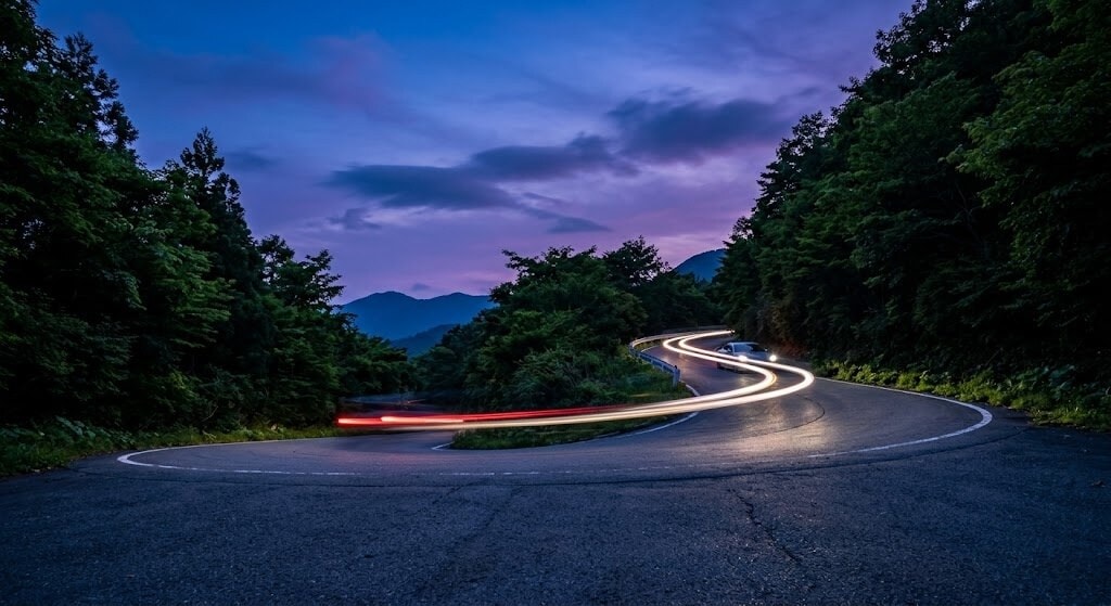 Winding mountain road at dusk, car light trails looping through a forested hillside.