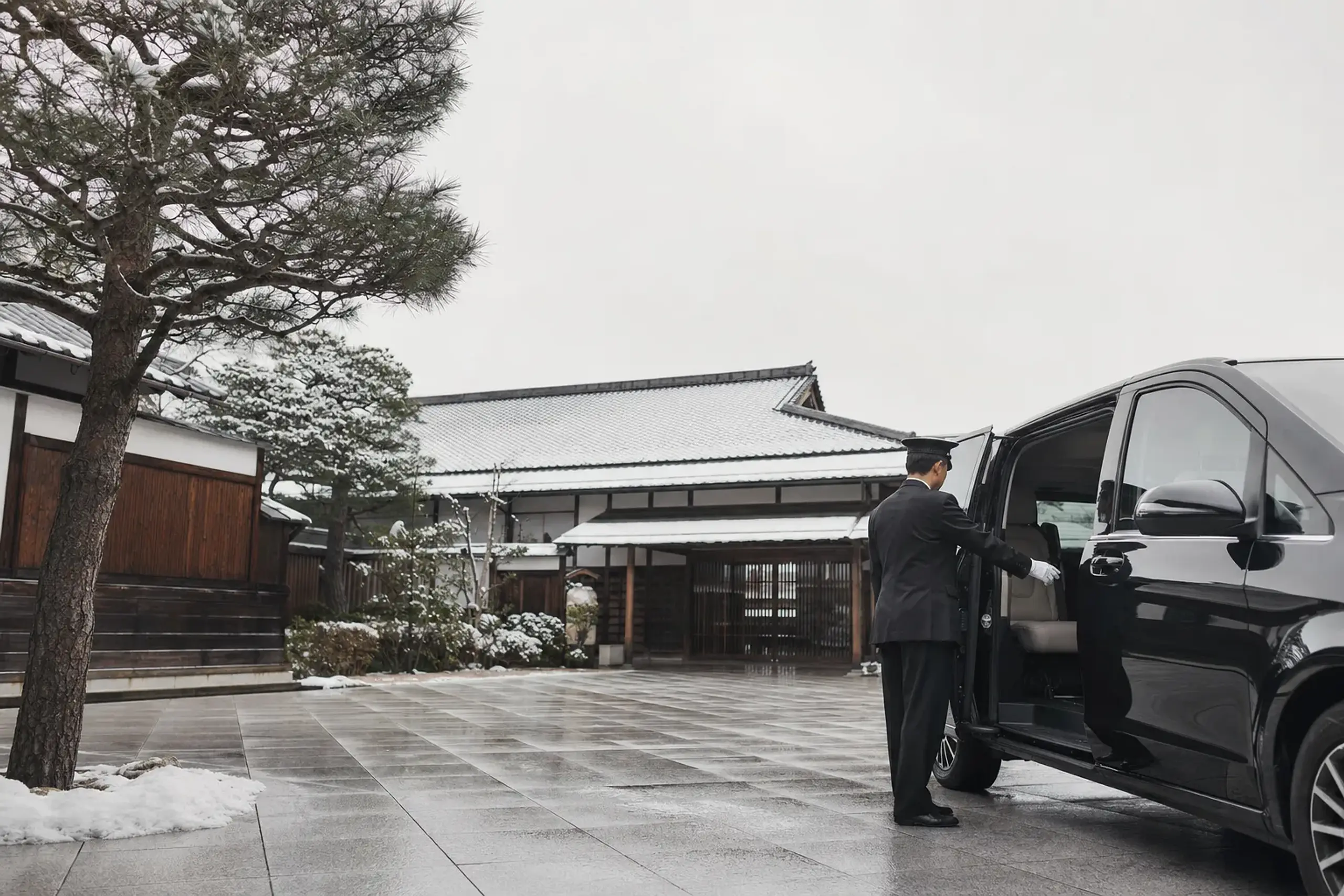 Chauffeur in suit opening the door of a black Mercedes passenger van in a traditional Japanese plaza setting.