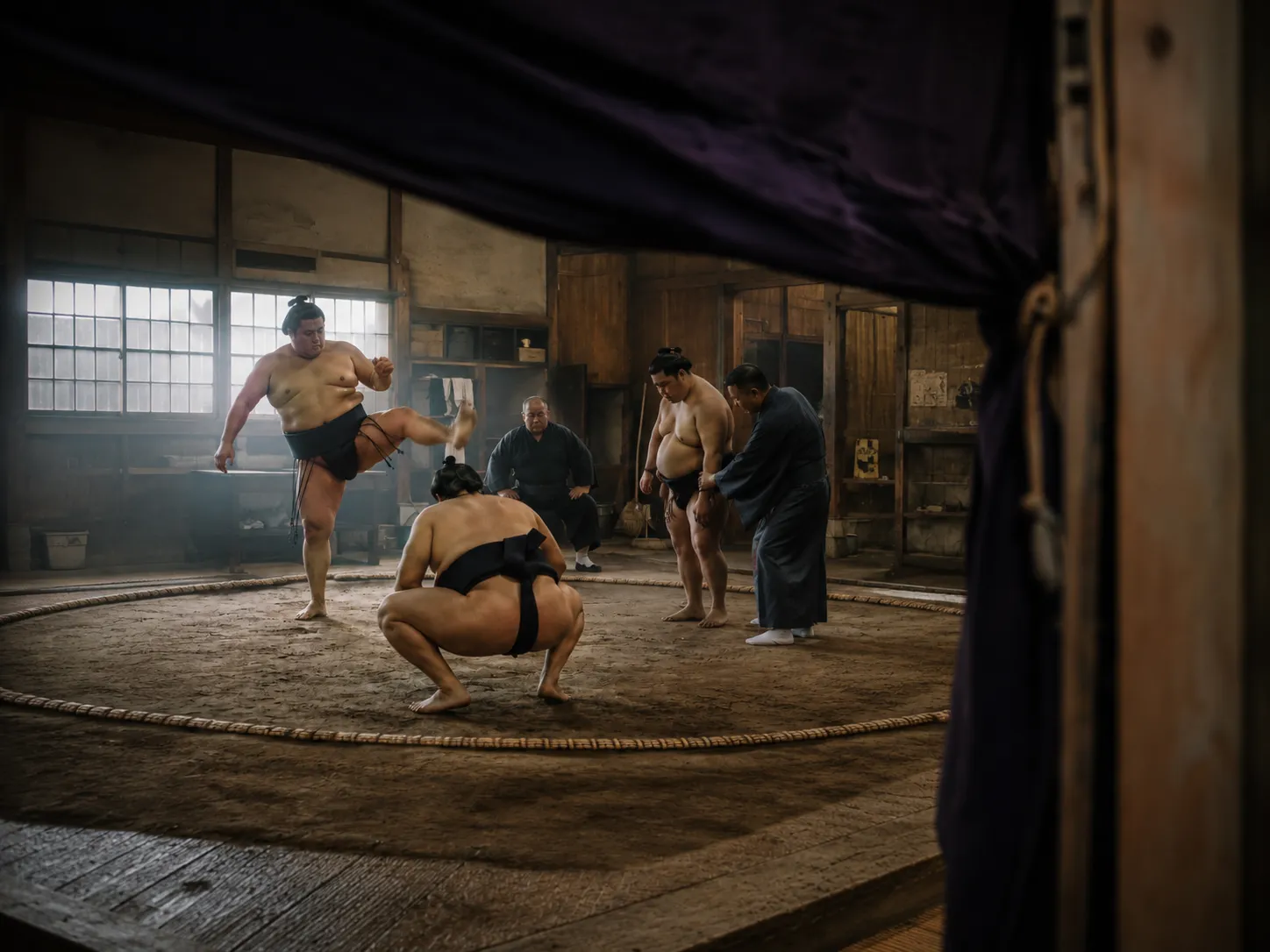 A sumo wrestler raising his leg in a shiko stomp during morning training while coaches and rikishi look on