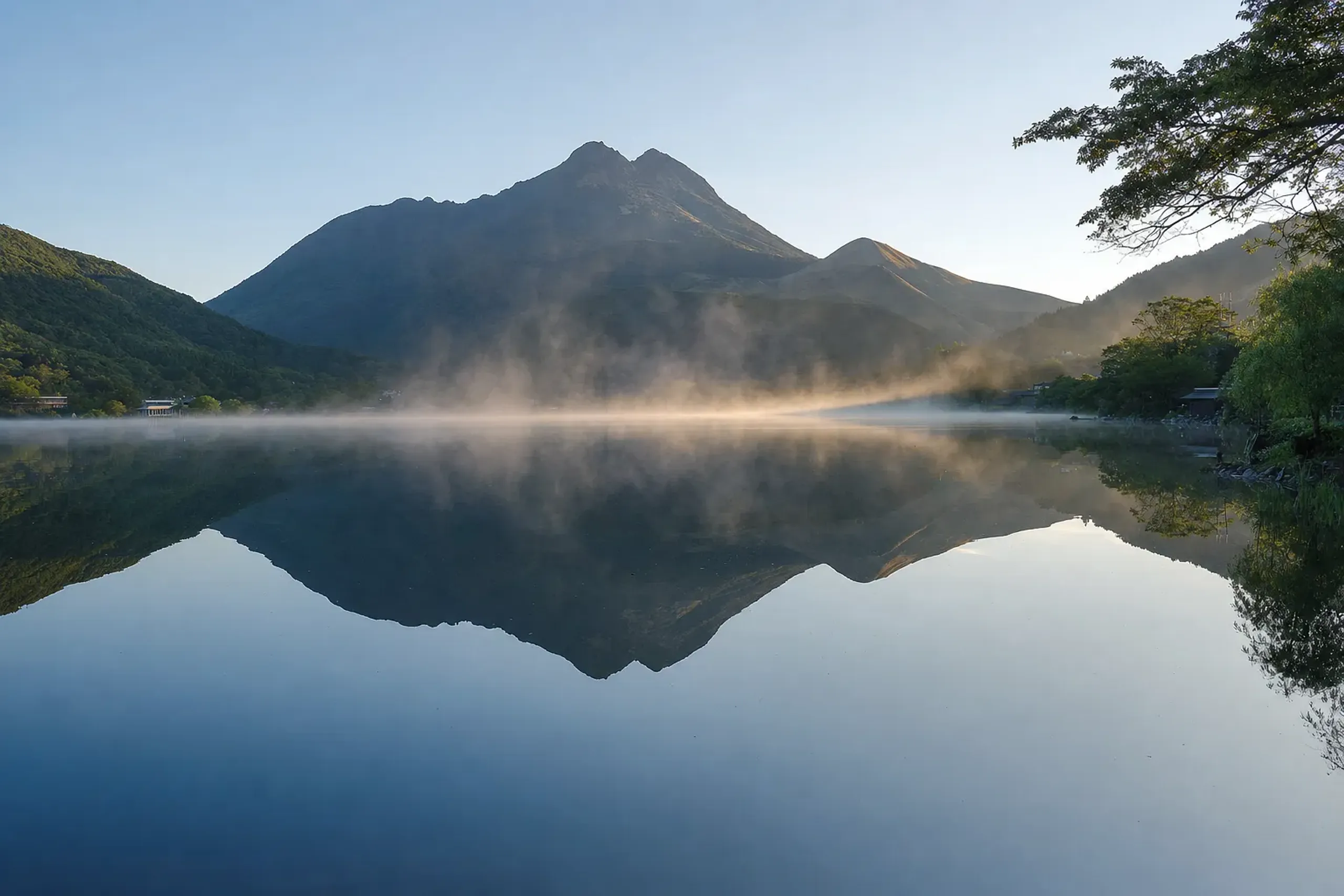 Steam rising off Kinrin Lake at dawn where cold spring and hot spring waters meet, with the twin-peaked silhouette of Mount Yufu reflected in the still surface