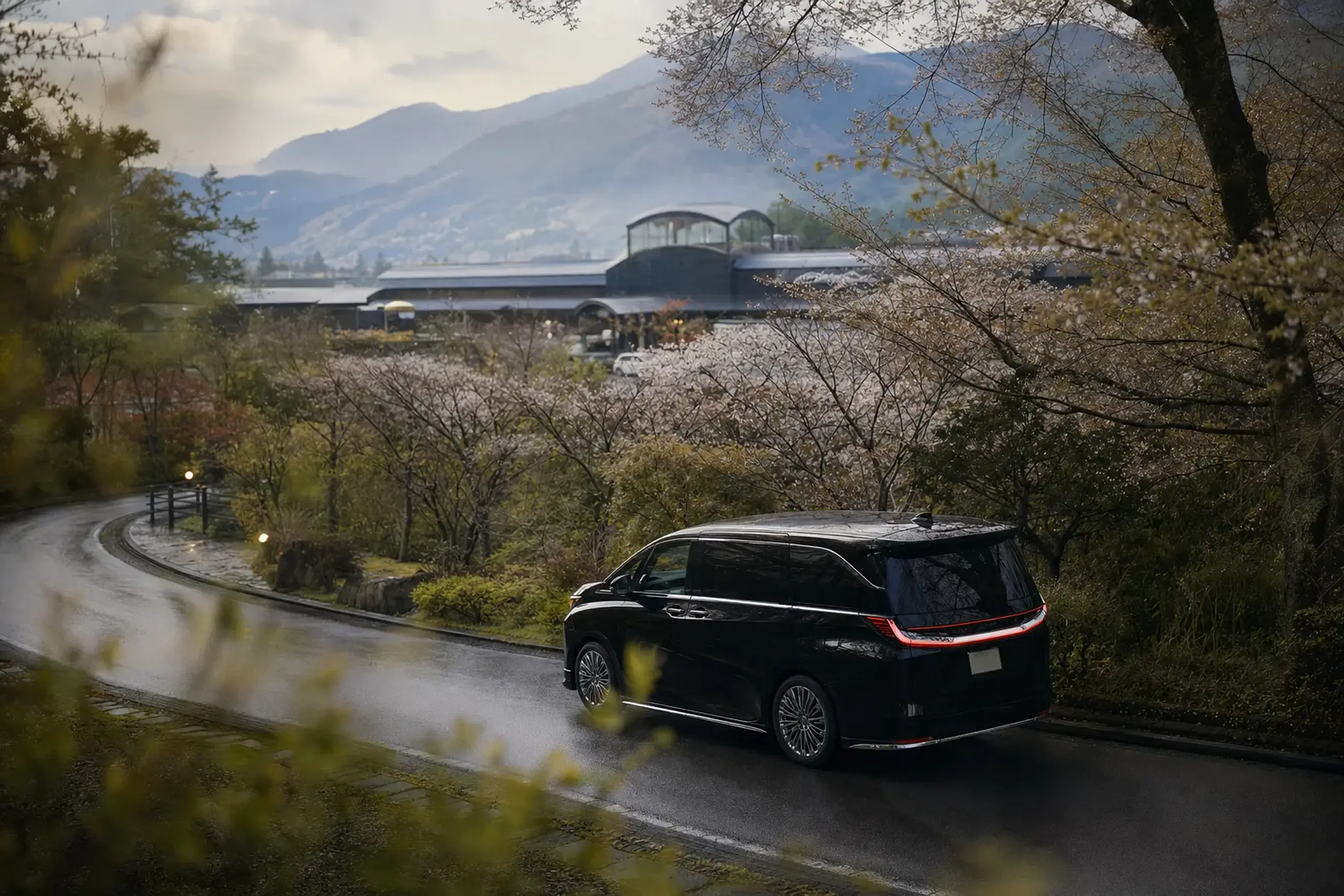 A Lexus LM 500 chauffeured transfer easing along the wooded hill road approaching JR Yufuin Station