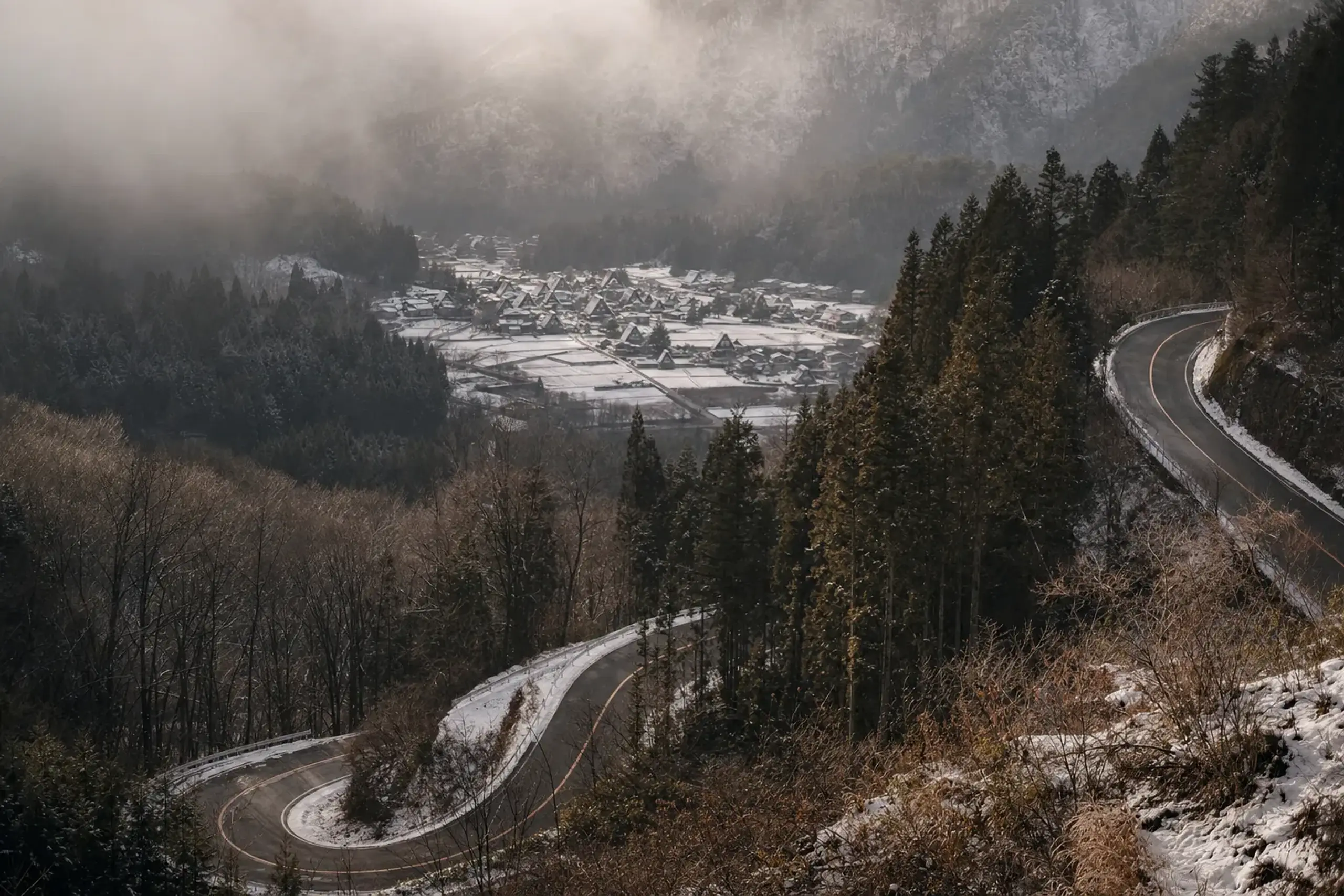 A winding mountain road descending through the steep forested valleys of the Japanese Alps toward the thatched-roof village of Shirakawa-go