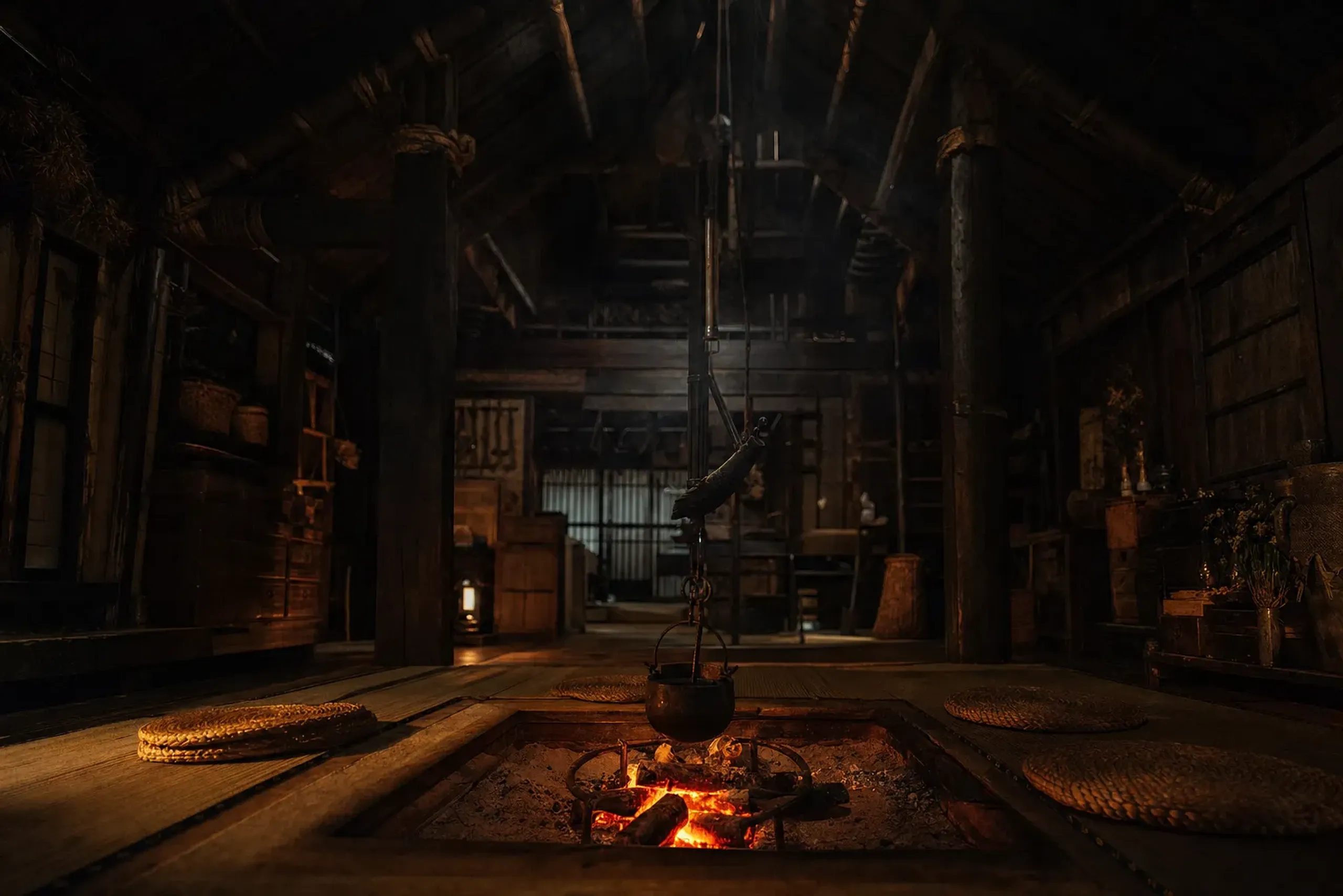 The smoke-blackened cedar interior of a gassho-zukuri farmhouse with a glowing open irori hearth on the ground floor and steep timber beams rising to the upper silkworm-cultivation floors