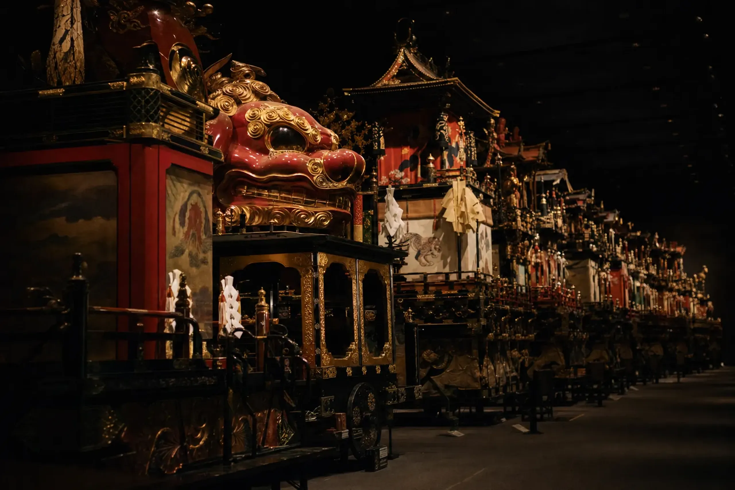 The fourteen Karatsu Kunchi hikiyama floats displayed indoors in a row at the Hikiyama Exhibition Hall, their lacquered forms lit under museum lighting