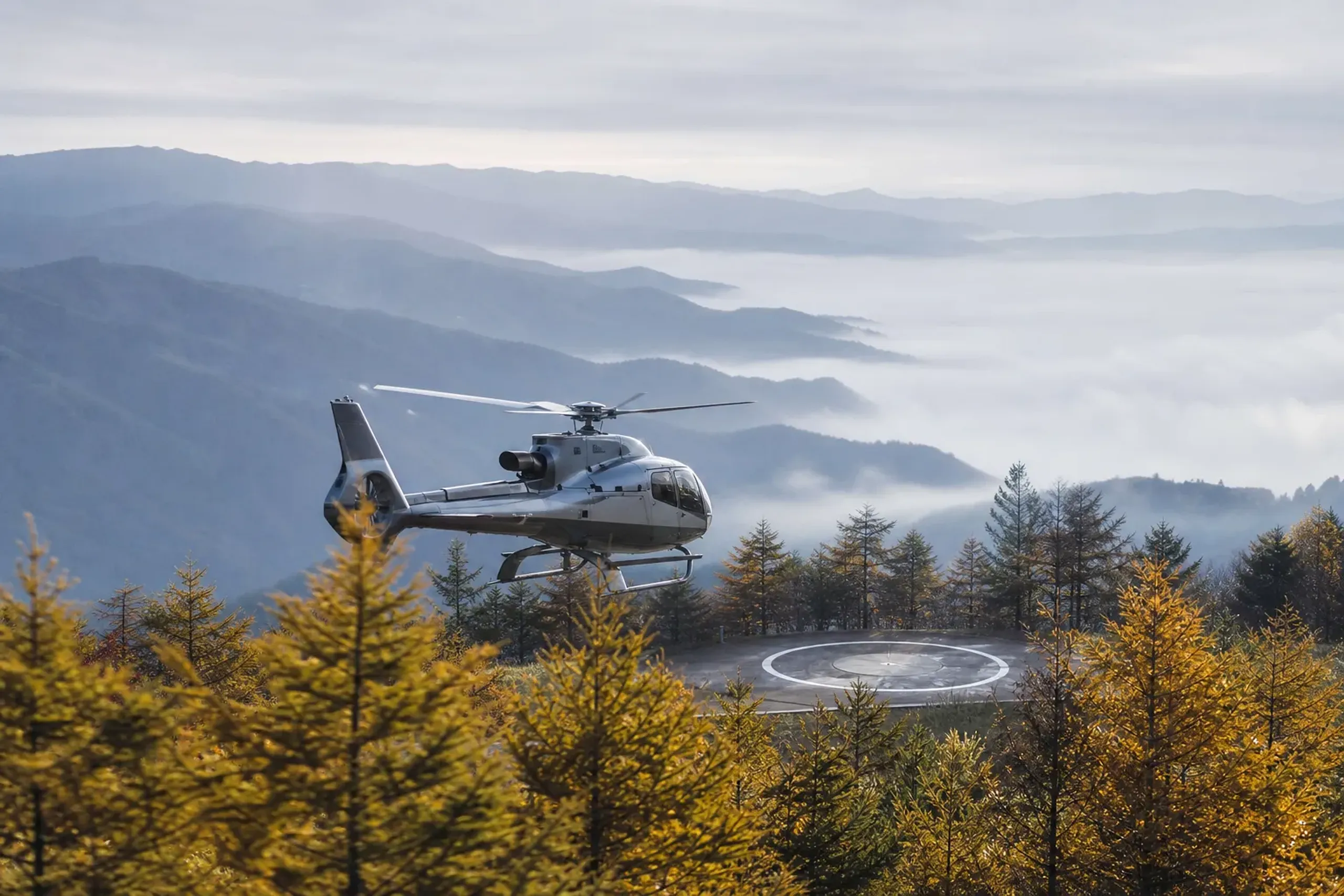 A private helicopter descending toward an alpine landing zone amid the Japanese larch forests of Karuizawa, with the misty Nagano highland ridgelines in the background