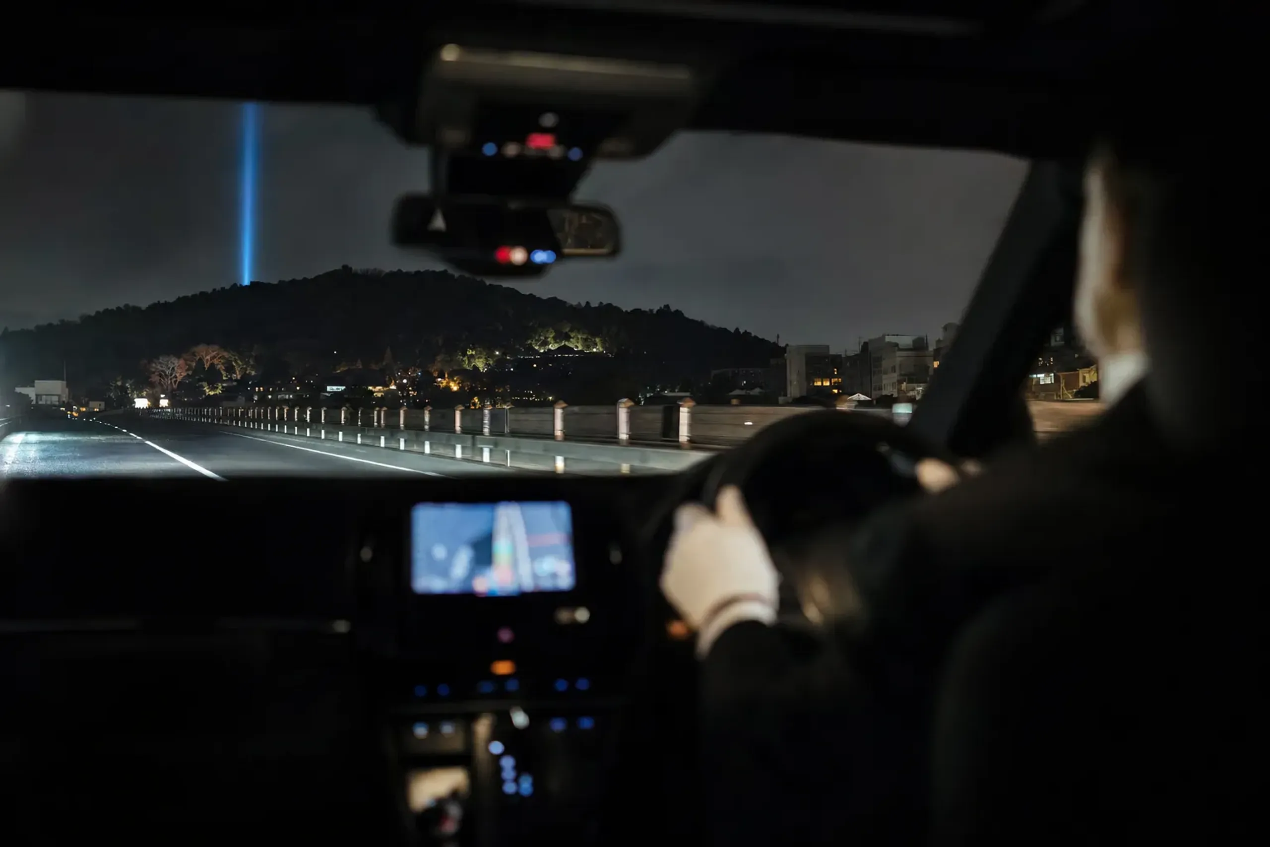A private car crossing the Kamo River at night with the distant blue Kannon Jihi-kō beam visible rising over Higashiyama
