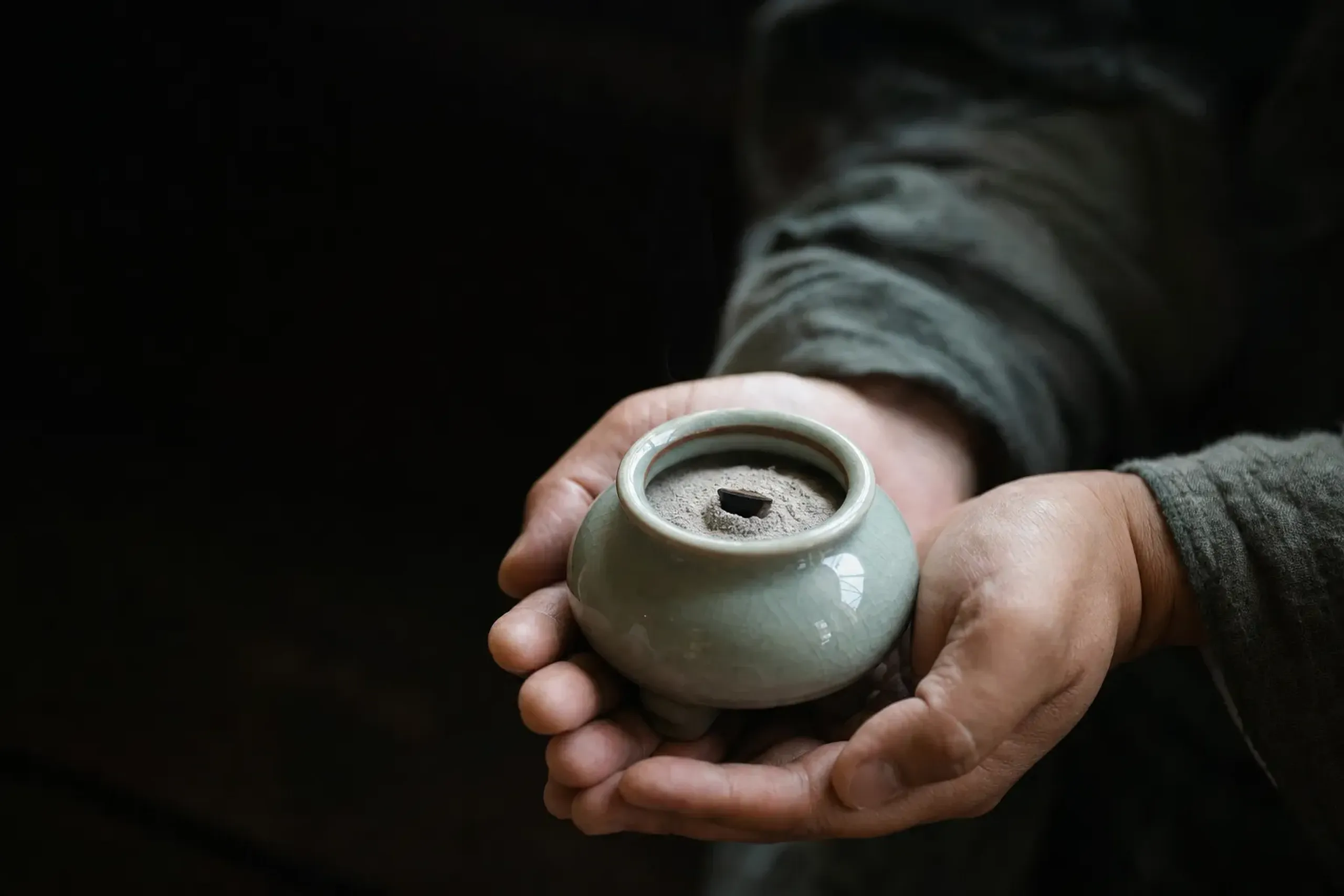 A small chip of jinkō agarwood warming over heated ash inside a porcelain kōro incense burner, thin heat rising with no smoke, held in cupped hands