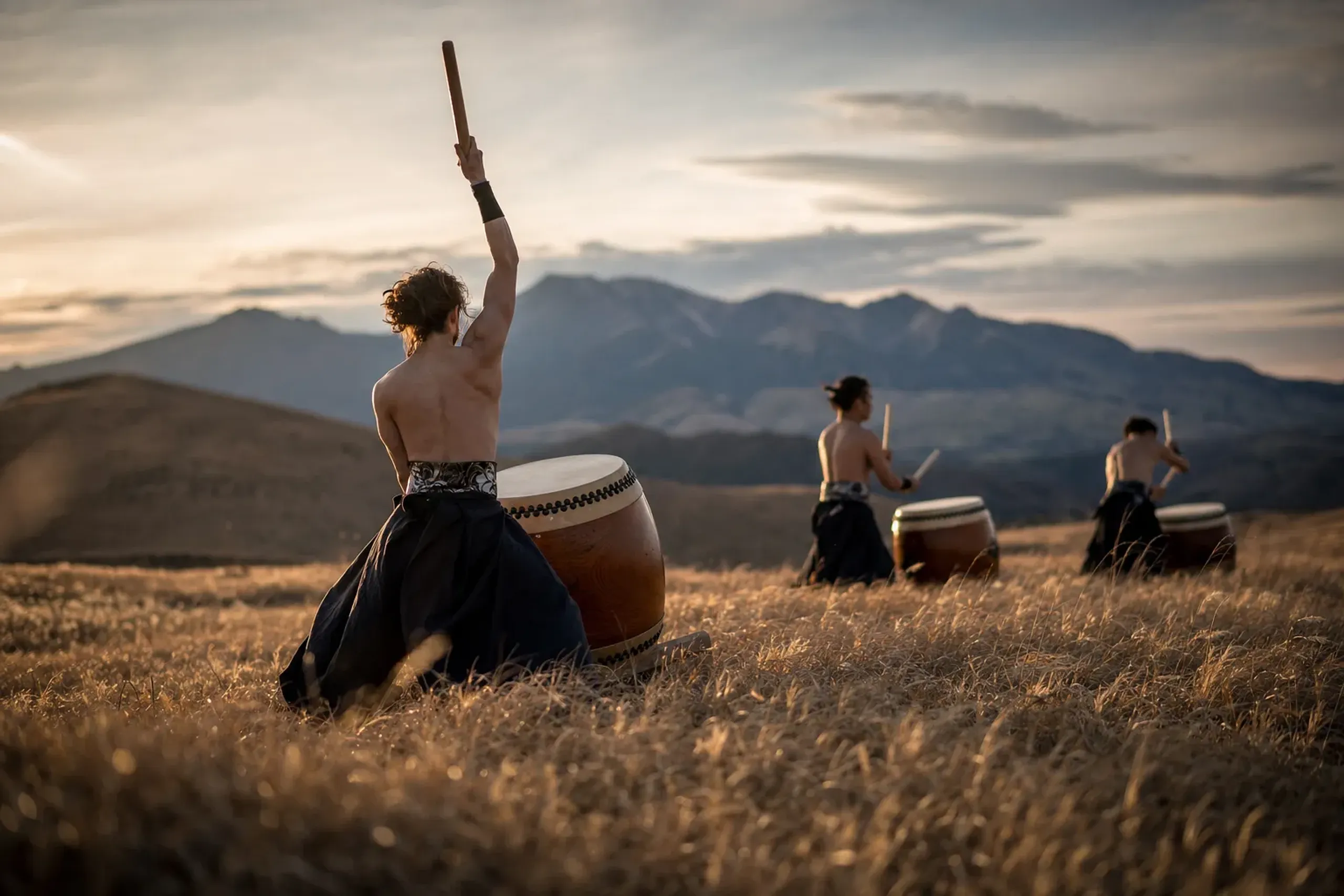 Taiko drummers of DRUM TAO performing at dusk on the volcanic grassland of the Kuju Plateau, mountains in silhouette behind them, golden low light at 1,000 metres elevation